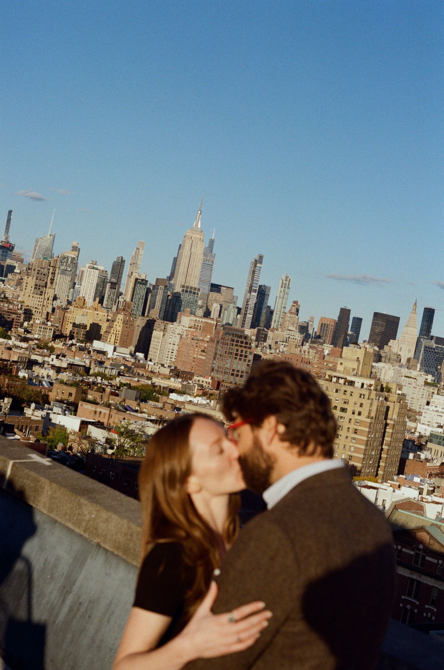 A couple shares a romantic kiss on a rooftop with the New York City skyline visible in the background.