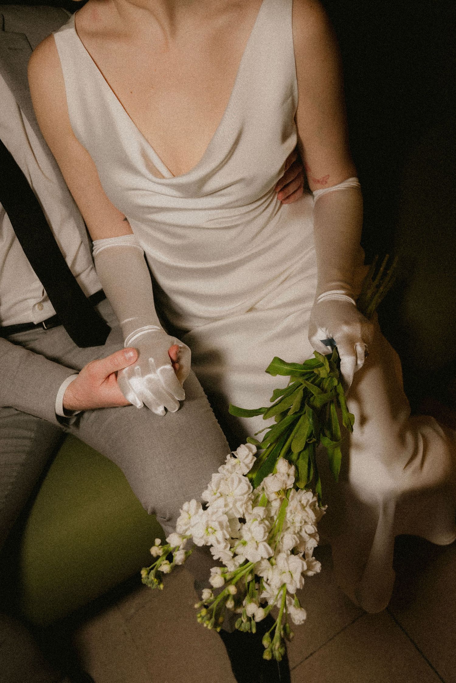 Bride in silk cowl neck gown holding white floral bouquet, wearing sheer gloves, seated beside groom in gray suit.