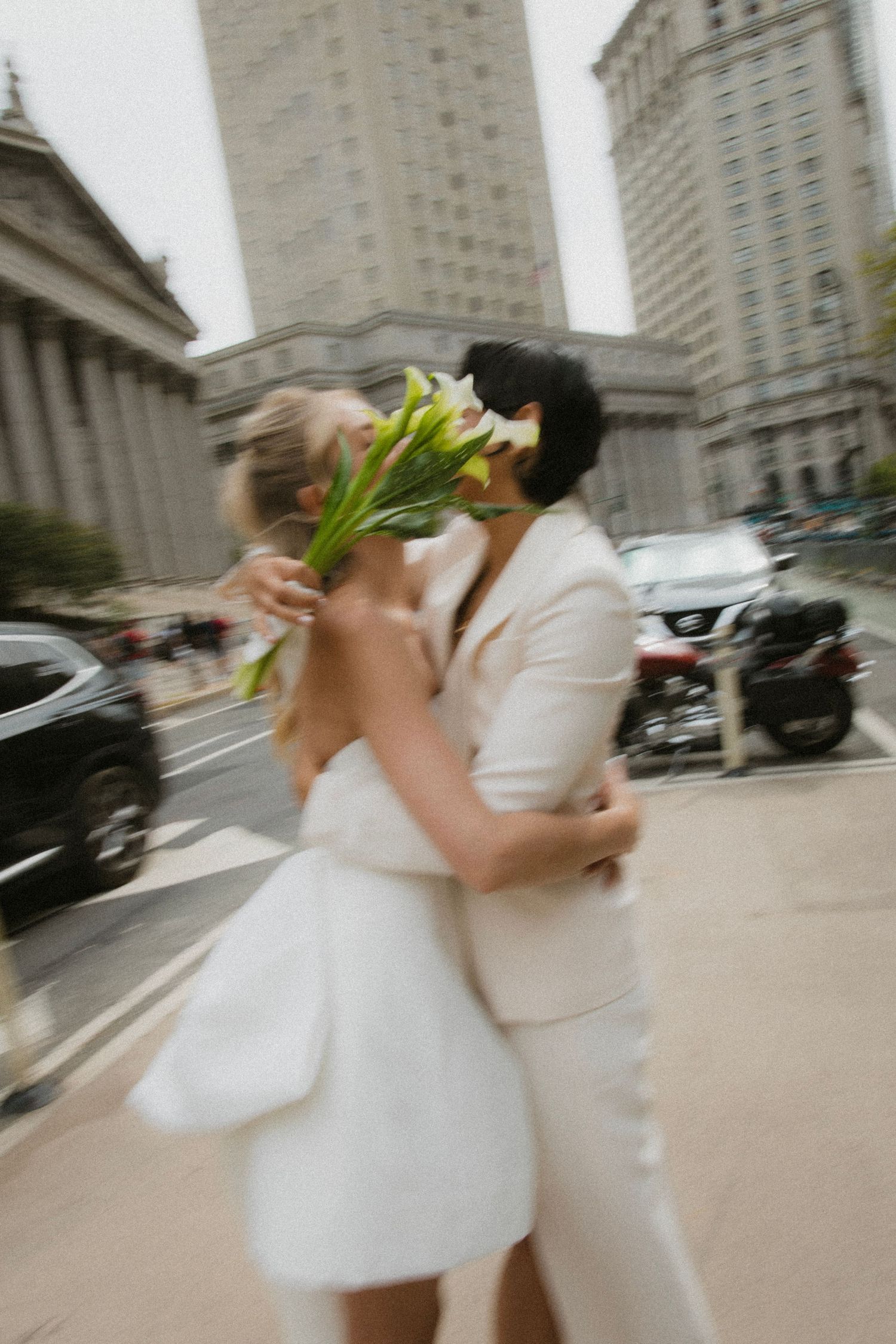 A couple in white wedding attire share a romantic kiss on a city street with tall buildings in the background.