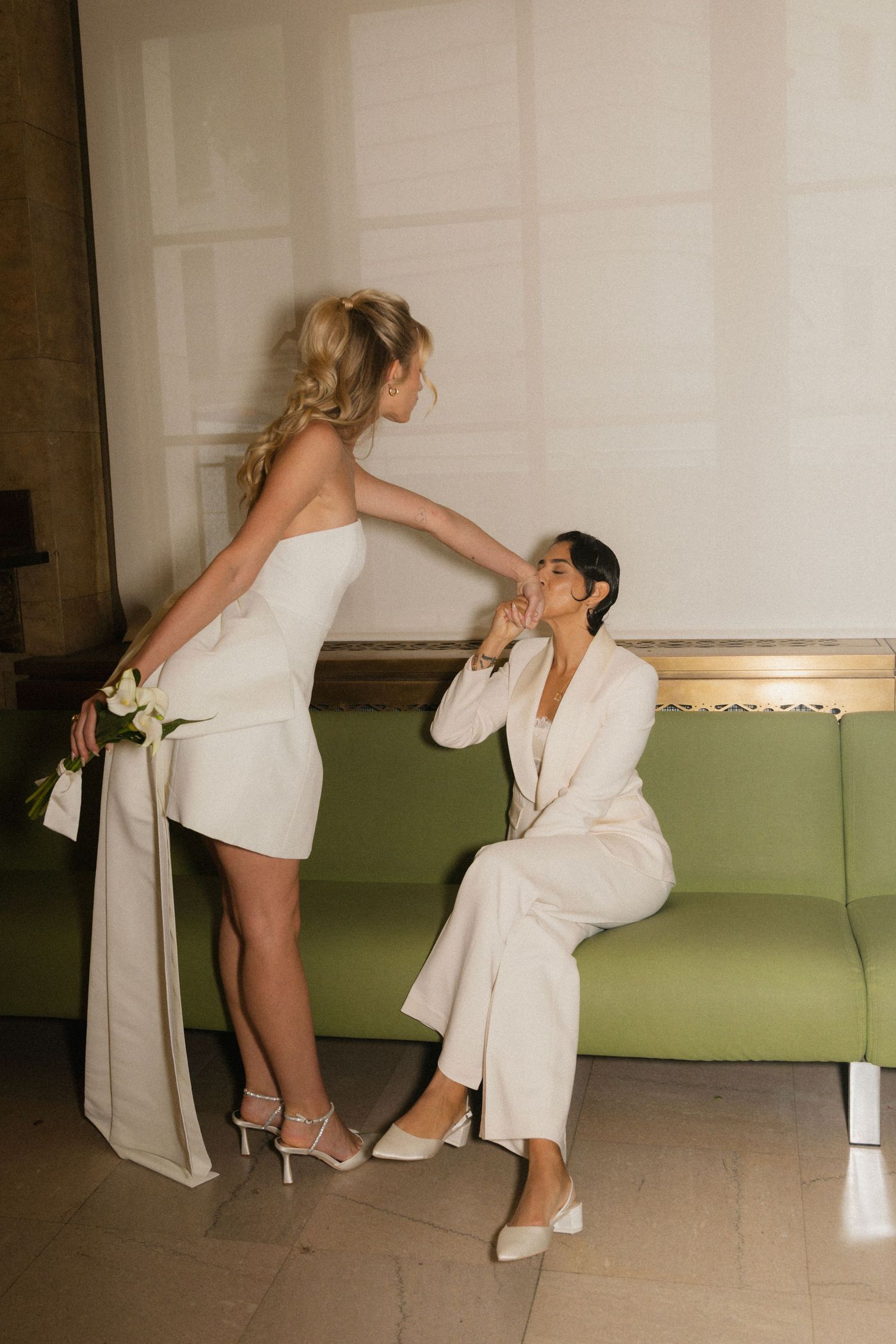 Two people in white outfits share an intimate moment on a green couch in an elegant indoor setting.