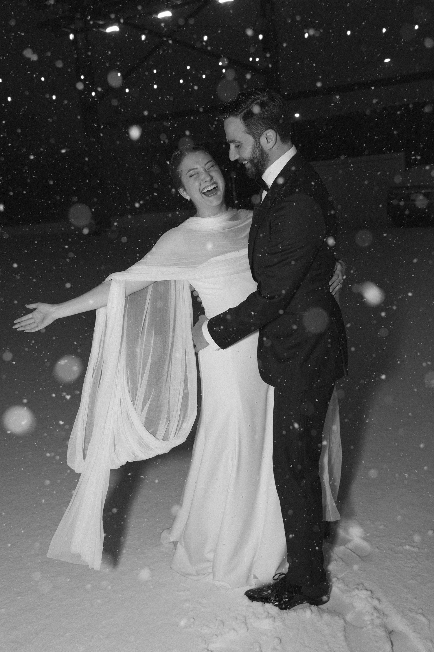 A joyful winter wedding scene with dancing couple in formal attire surrounded by falling snow.