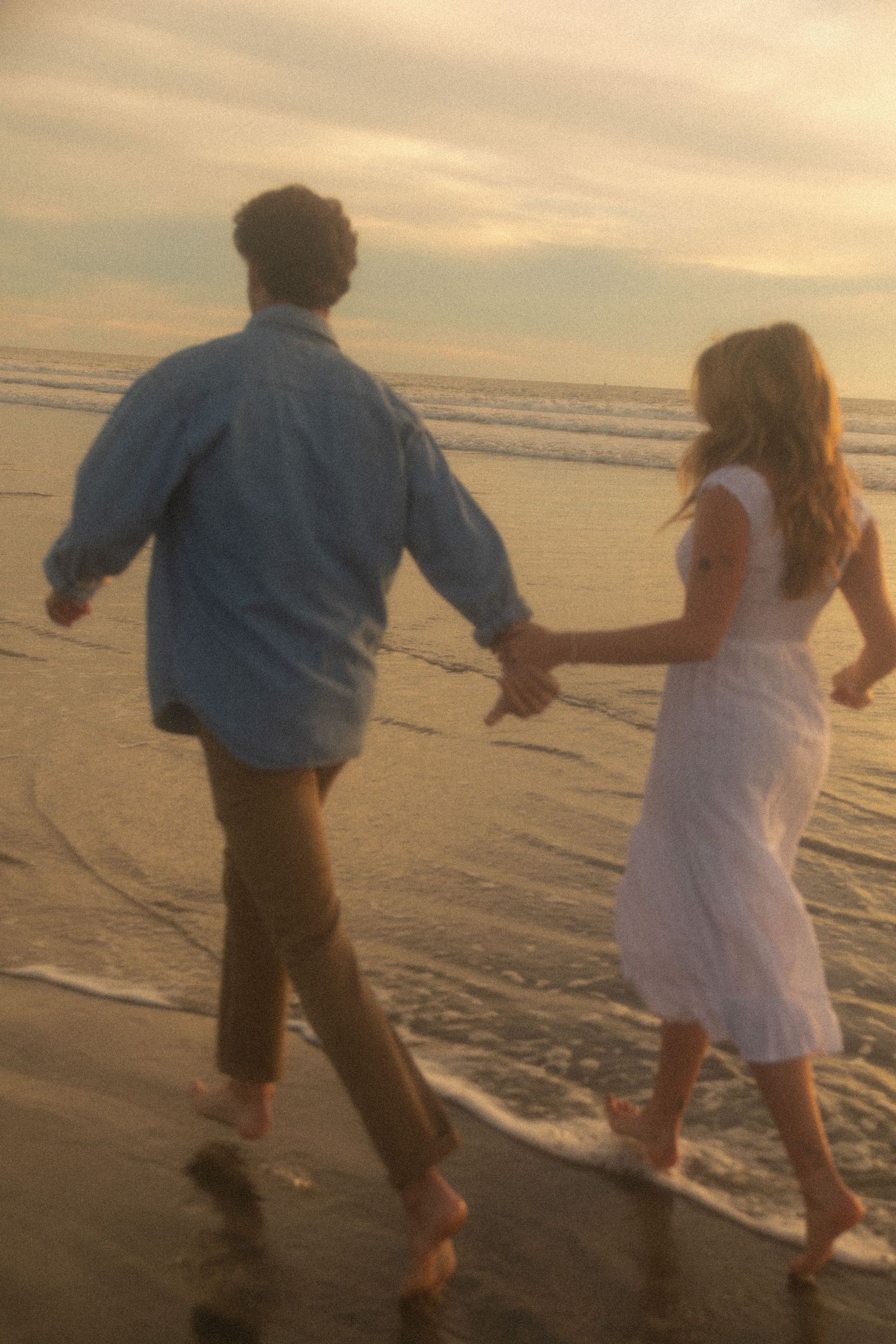 A couple runs playfully along the beach at sunset holding hands as waves lap at their feet.