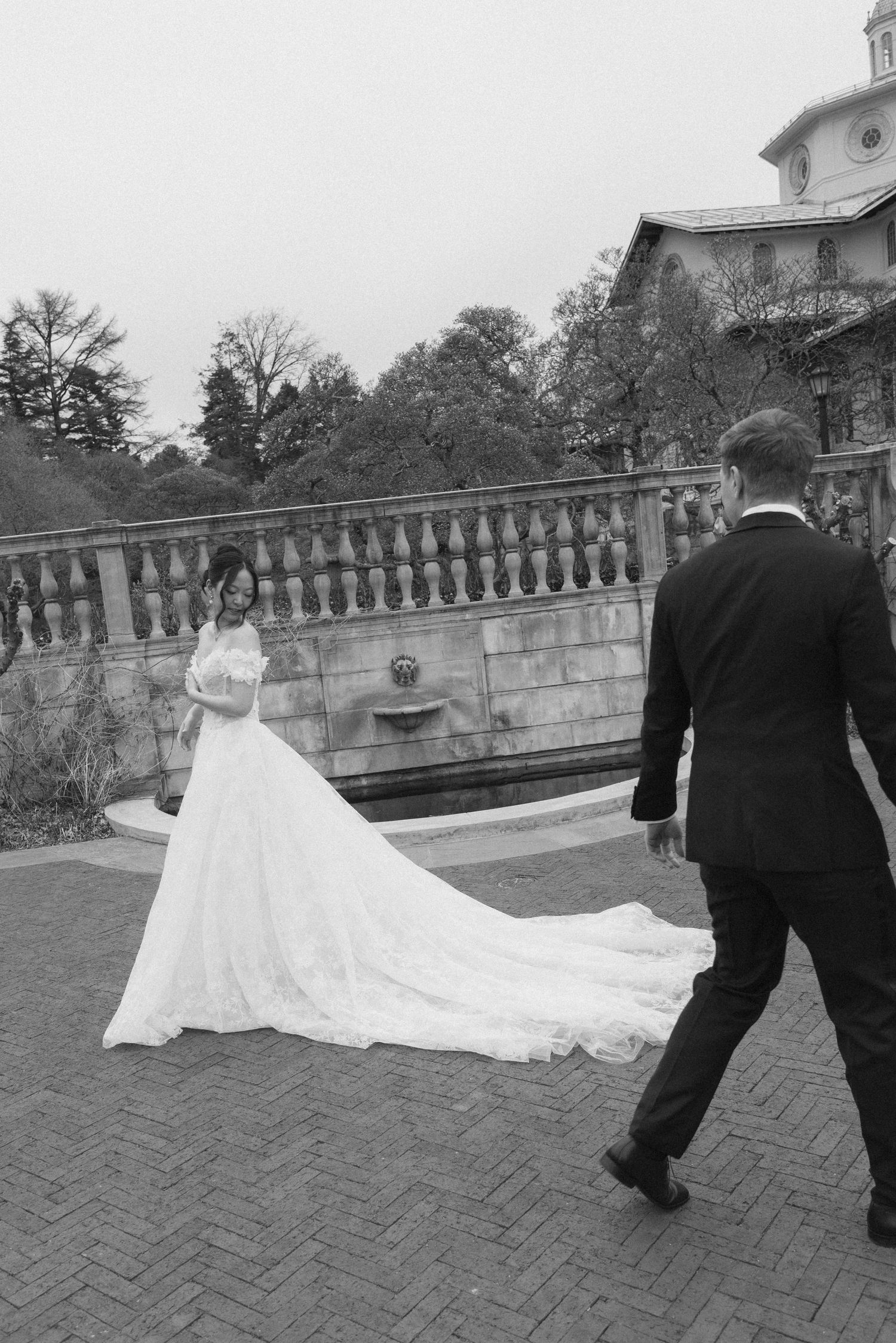 A bride in a flowing white dress with a long train walks across a brick patio with a man in a black suit.