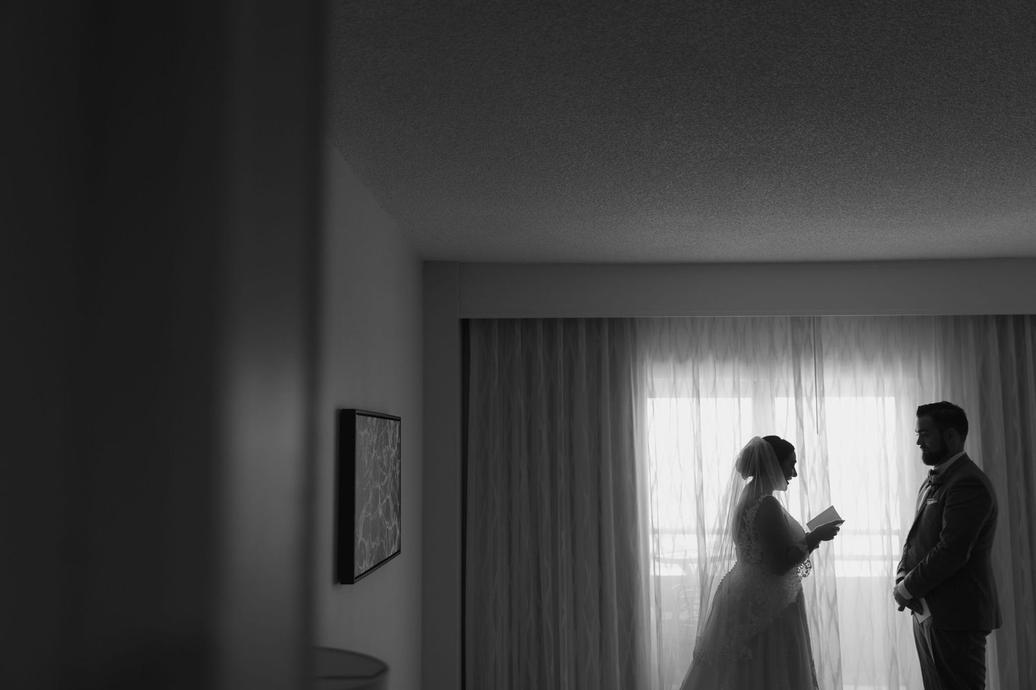 A silhouetted wedding scene in a bright hotel room with sunlight streaming through sheer curtains.
