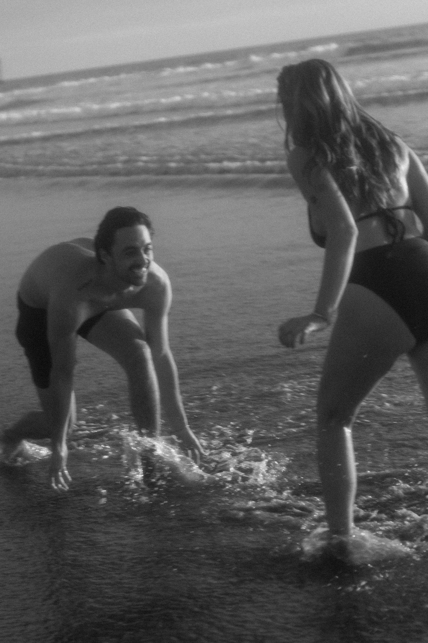 Sequential black and white frames show people splashing and playing in shallow ocean waves along a beach shoreline.