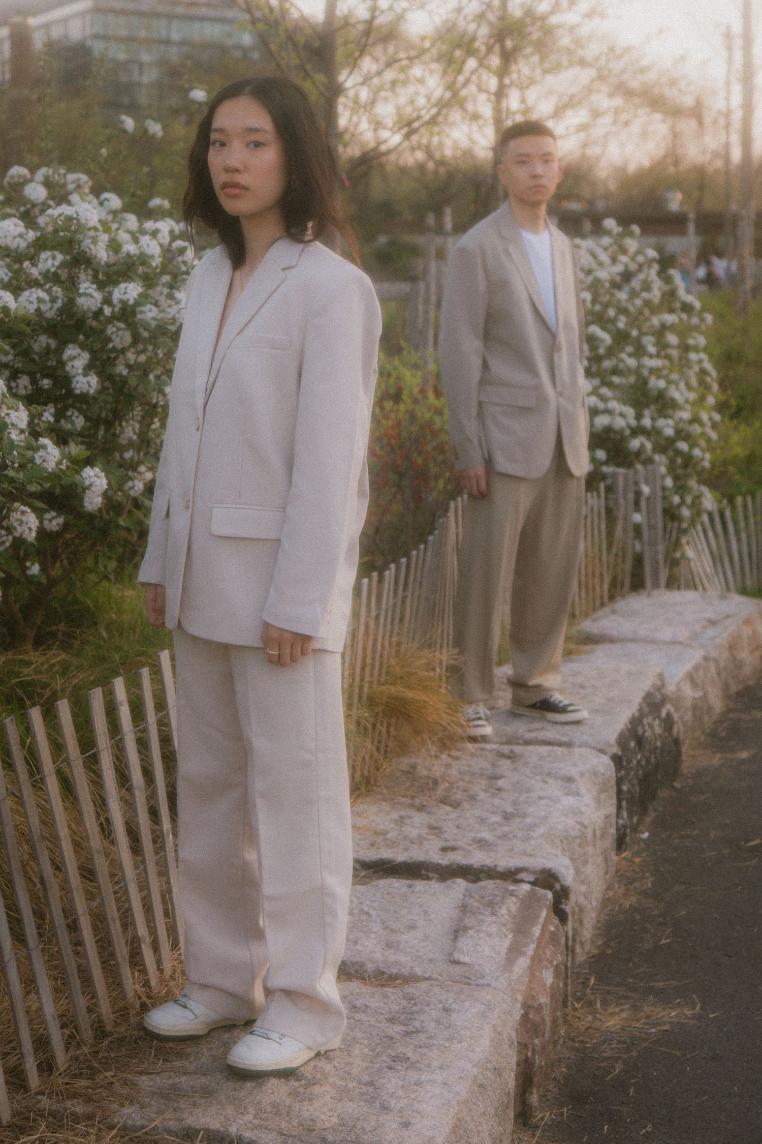 Two people in light colored suits stand on a stone ledge near white flowering bushes at sunset.