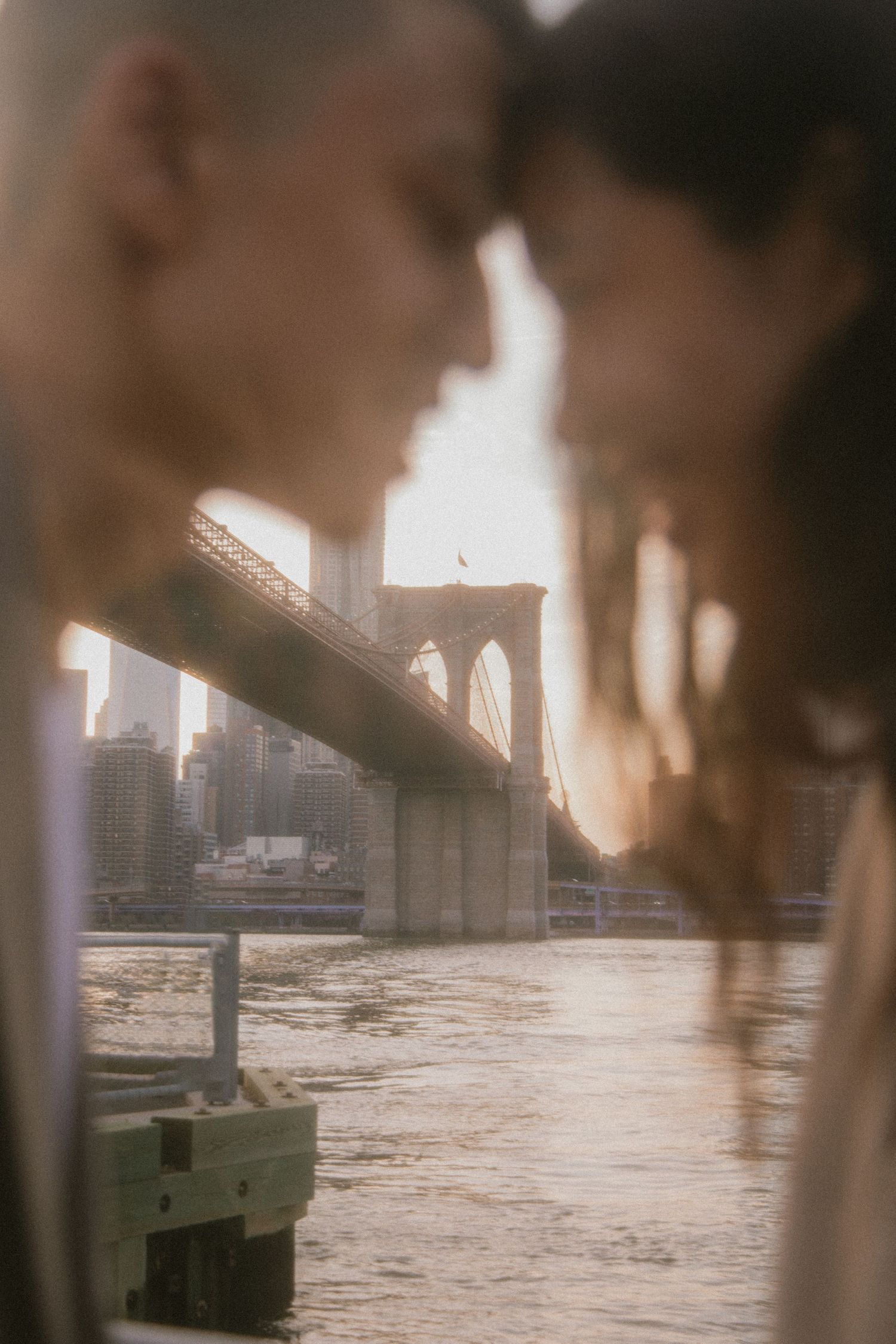Soft focus romantic portrait near Brooklyn Bridge at sunset with river and cityscape in background.
