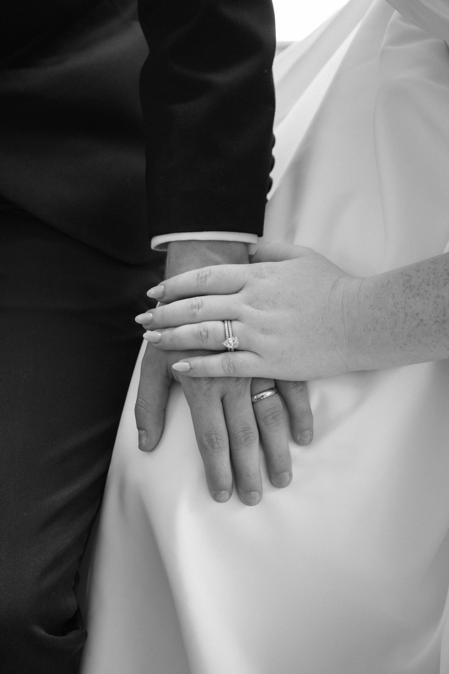 Close-up black and white photo of hands with wedding rings resting on formal attire.