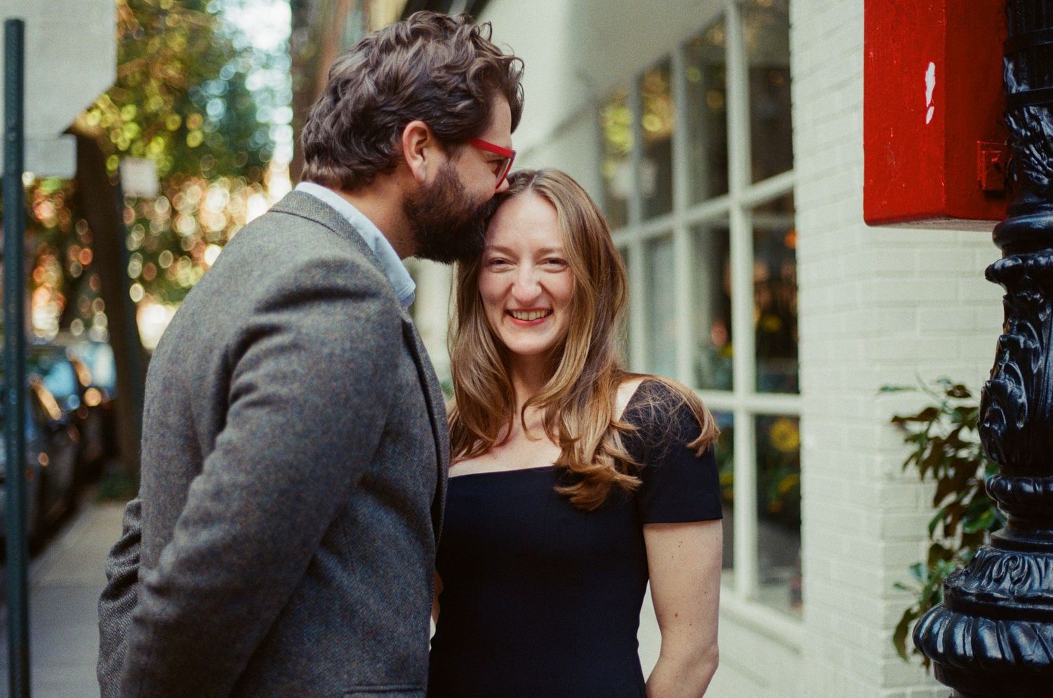 A couple shares a romantic moment together in an outdoor setting with sunlit trees and buildings in the background.