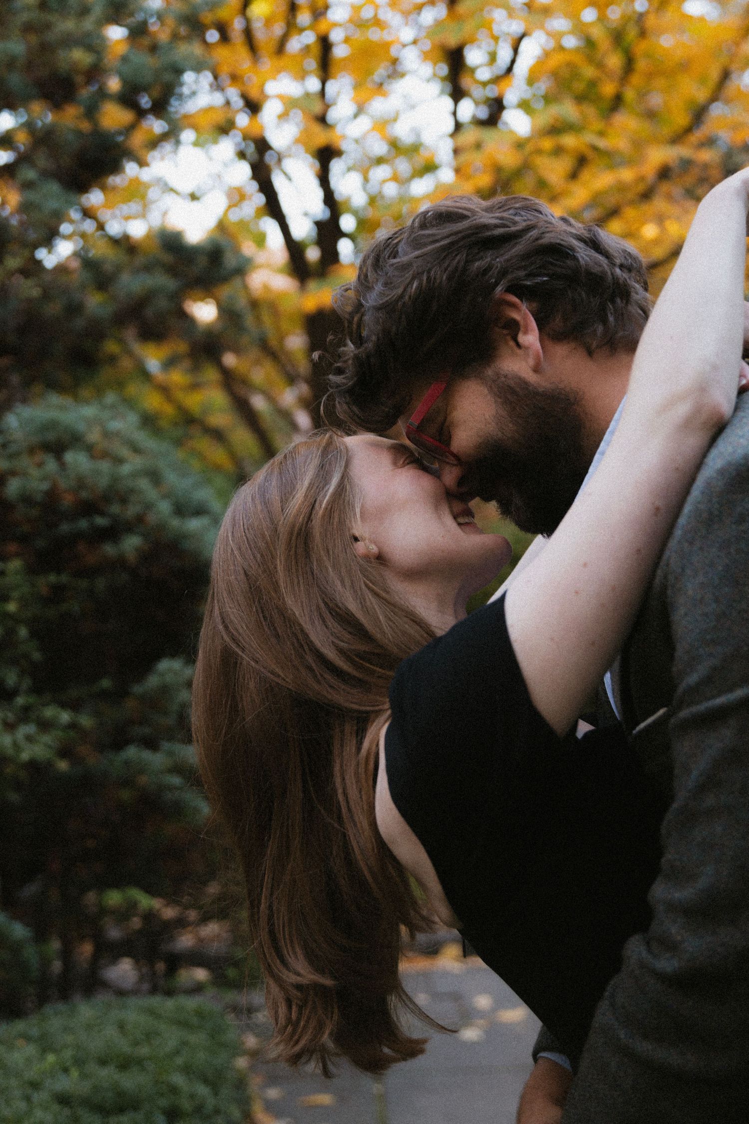 A romantic couple shares a tender kiss in a park with golden autumn foliage in the background.