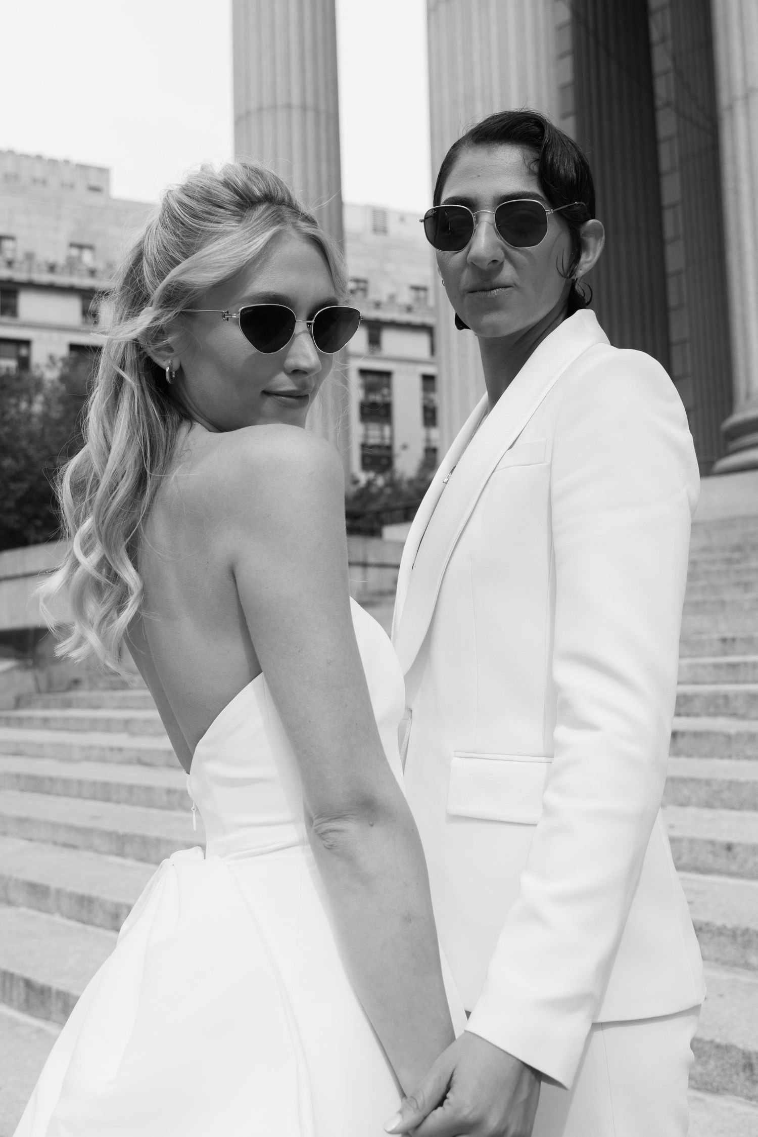 A couple in white attire and sunglasses poses together on stone steps near classical columns.