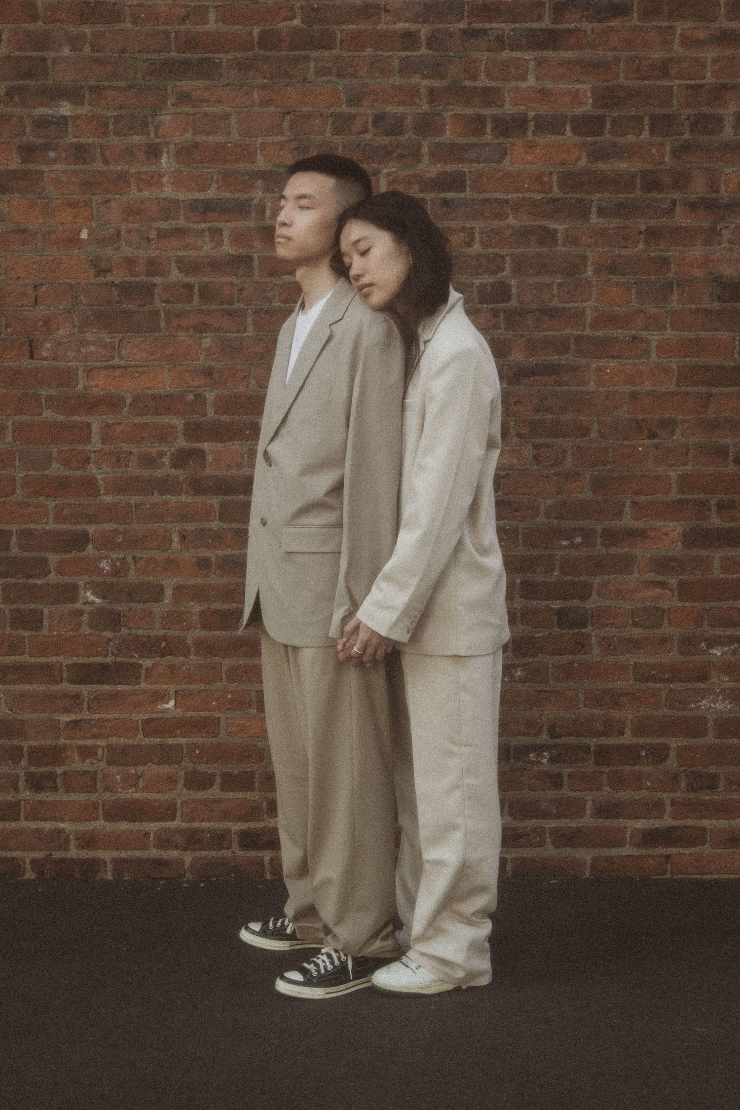 Two figures in coordinating neutral suits lean against a brick wall while wearing casual sneakers.