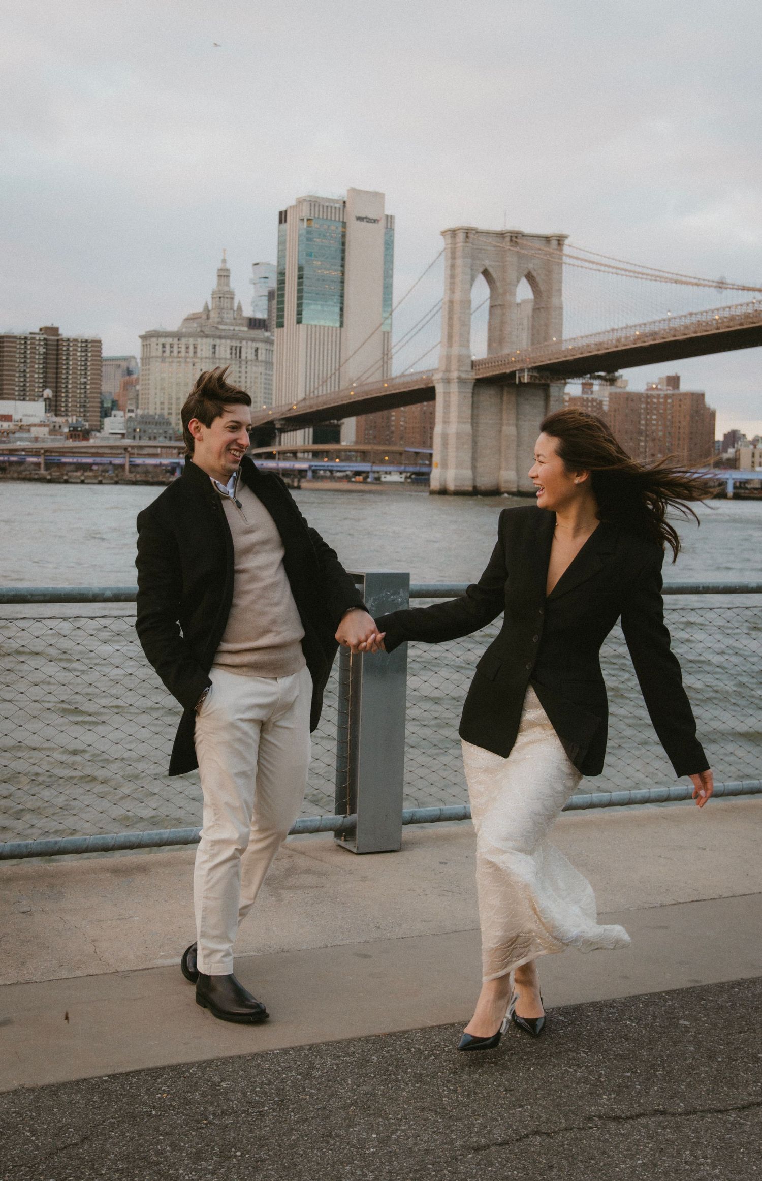 Two people dancing and laughing together on a waterfront promenade with the Brooklyn Bridge in the background.