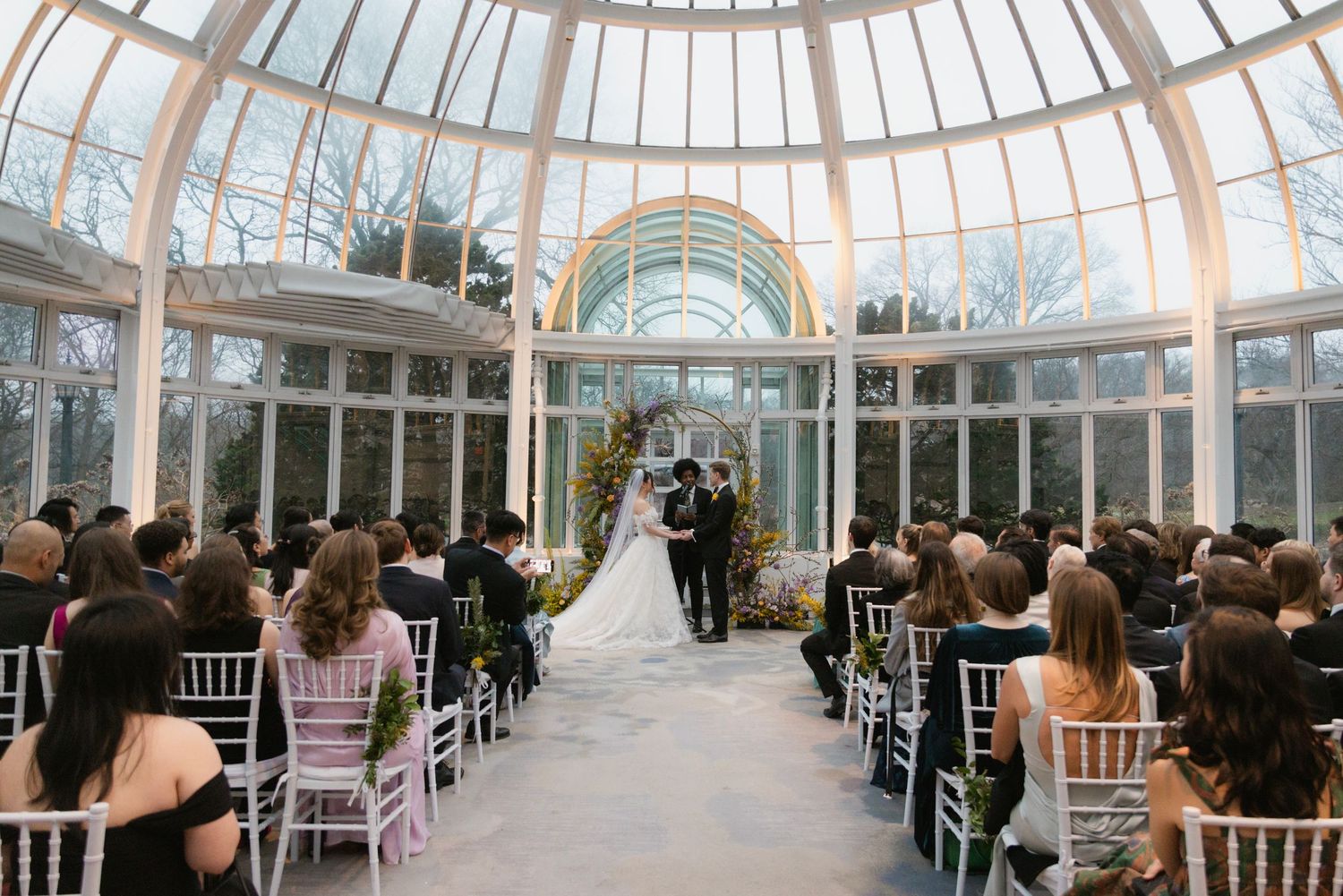 Indoor wedding ceremony in elegant glass conservatory with guests seated.
