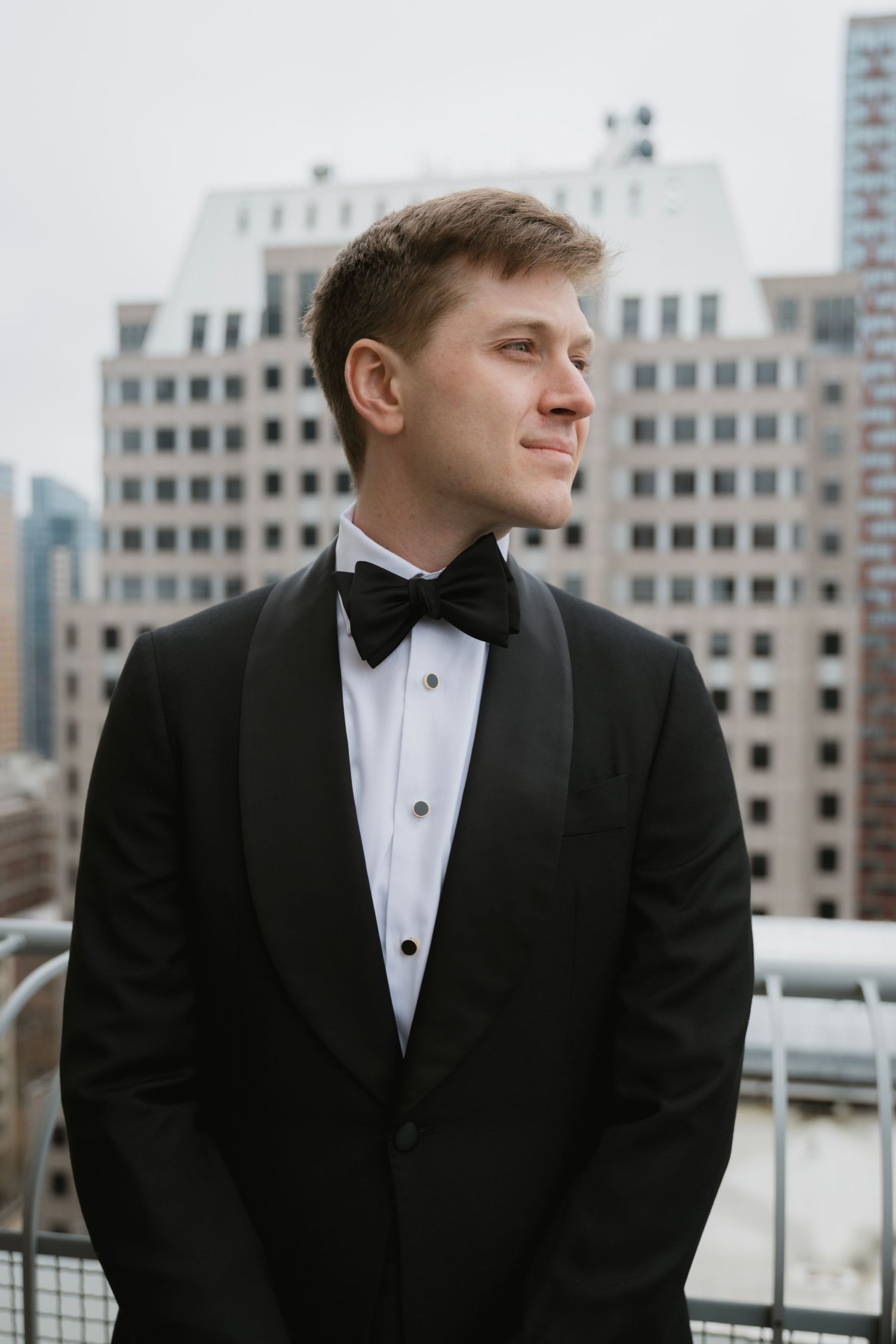 Groom in tuxedo standing against urban backdrop with tall buildings.