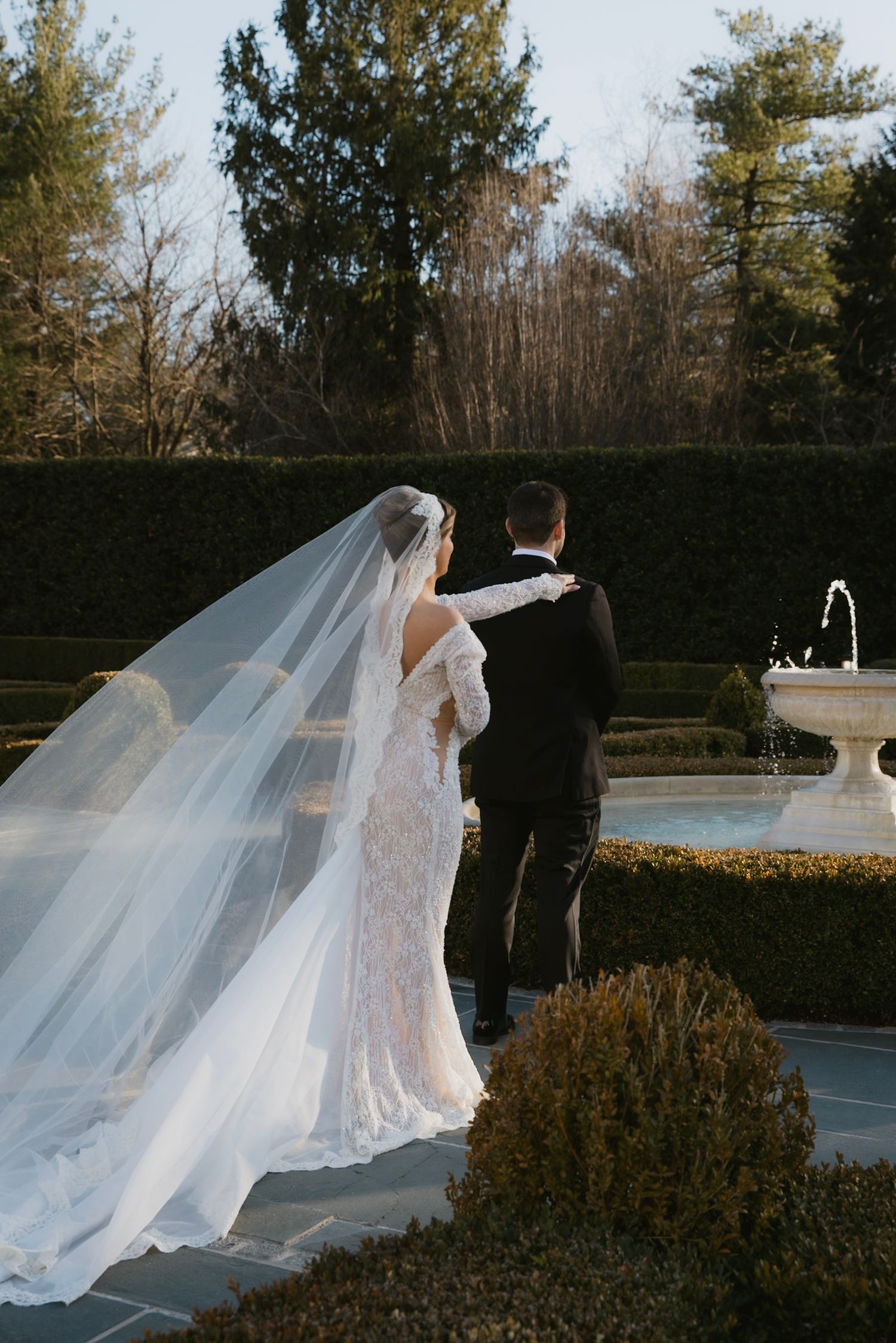 Romantic garden wedding photo with flowing veil and fountain in the background.