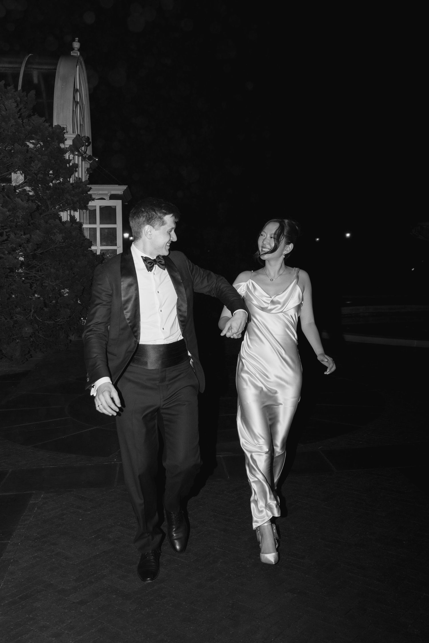 A couple in elegant evening wear walks together at night, with the person in a tuxedo and their partner in a silk dress.