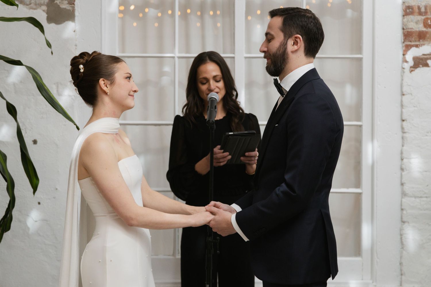 An intimate wedding ceremony takes place in a bright industrial space with white brick walls and string lights.