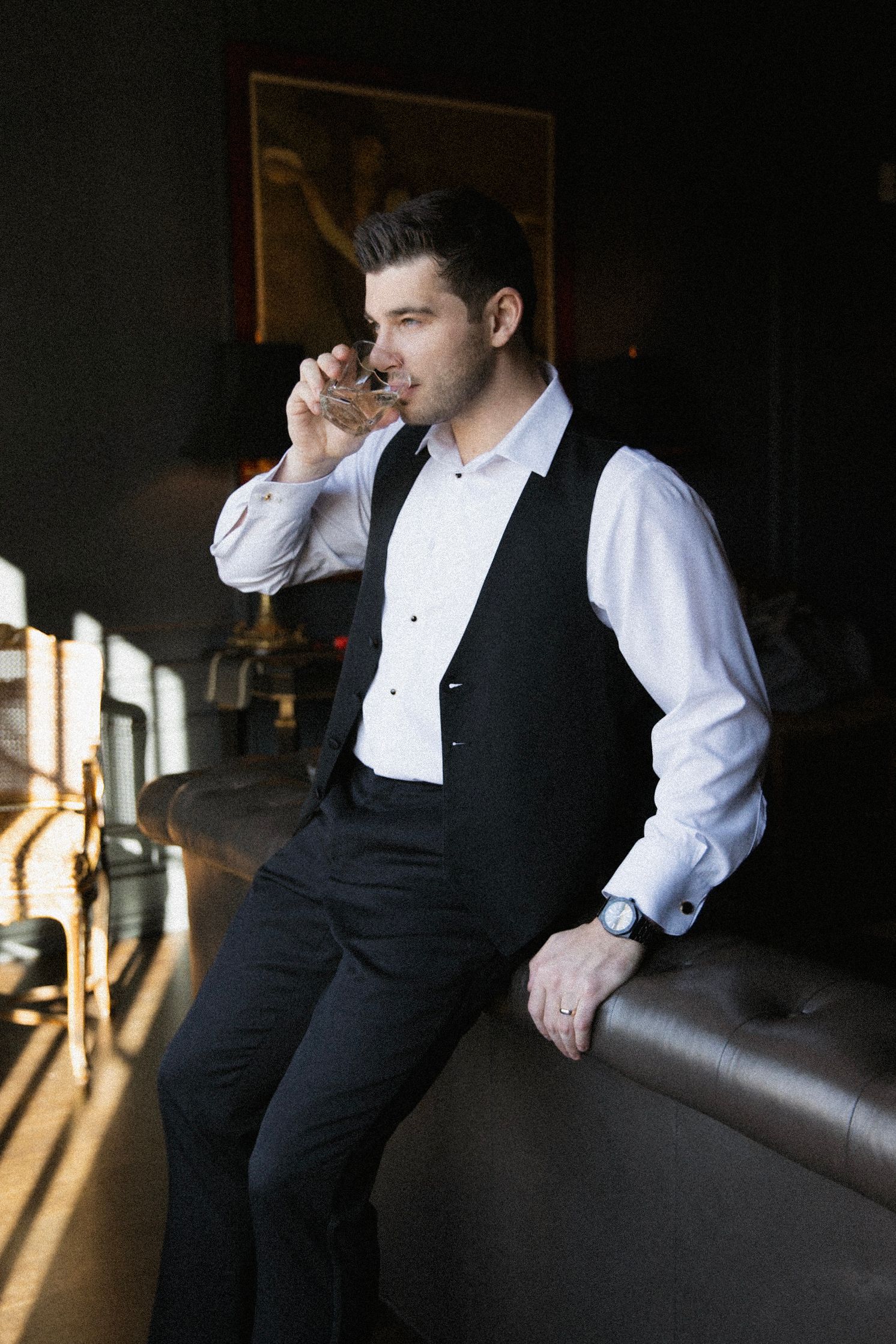 A person in formal evening attire sips a drink while leaning against a leather couch in moody lighting.