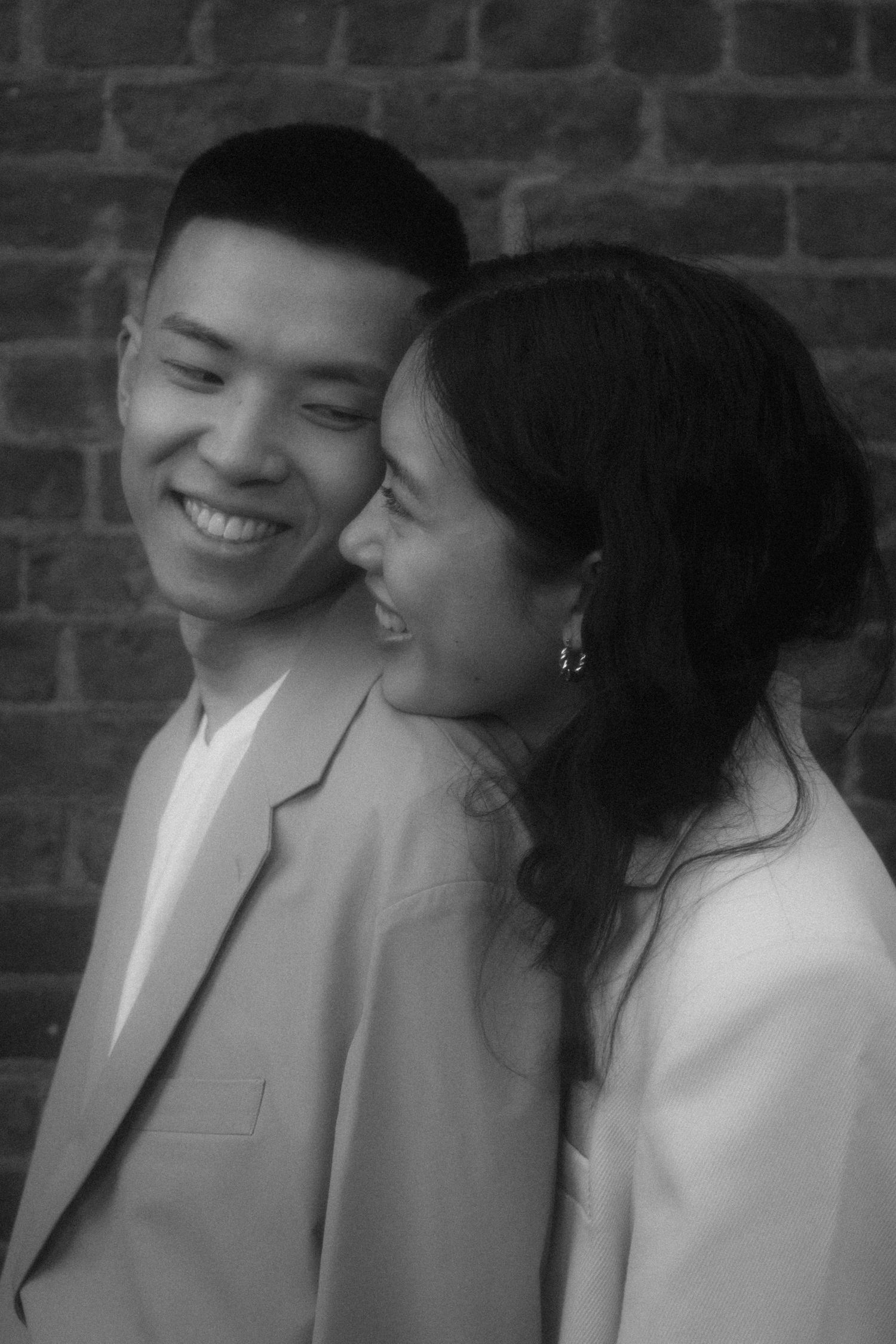 A joyful black and white portrait of a couple smiling together against a brick wall.