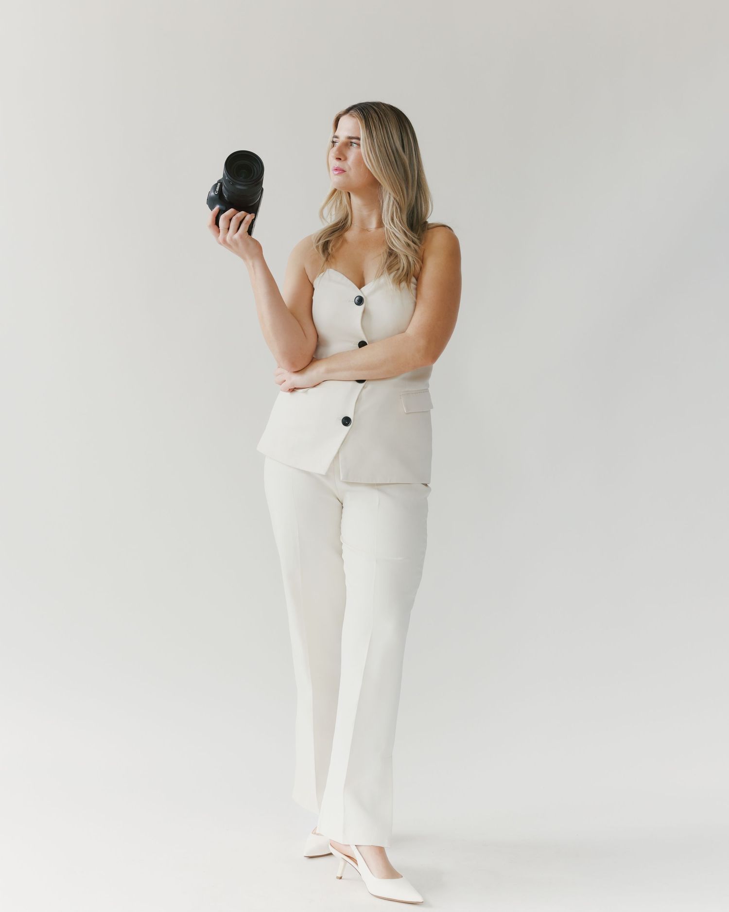 Portrait of a woman in white outfit holding camera against minimal background.