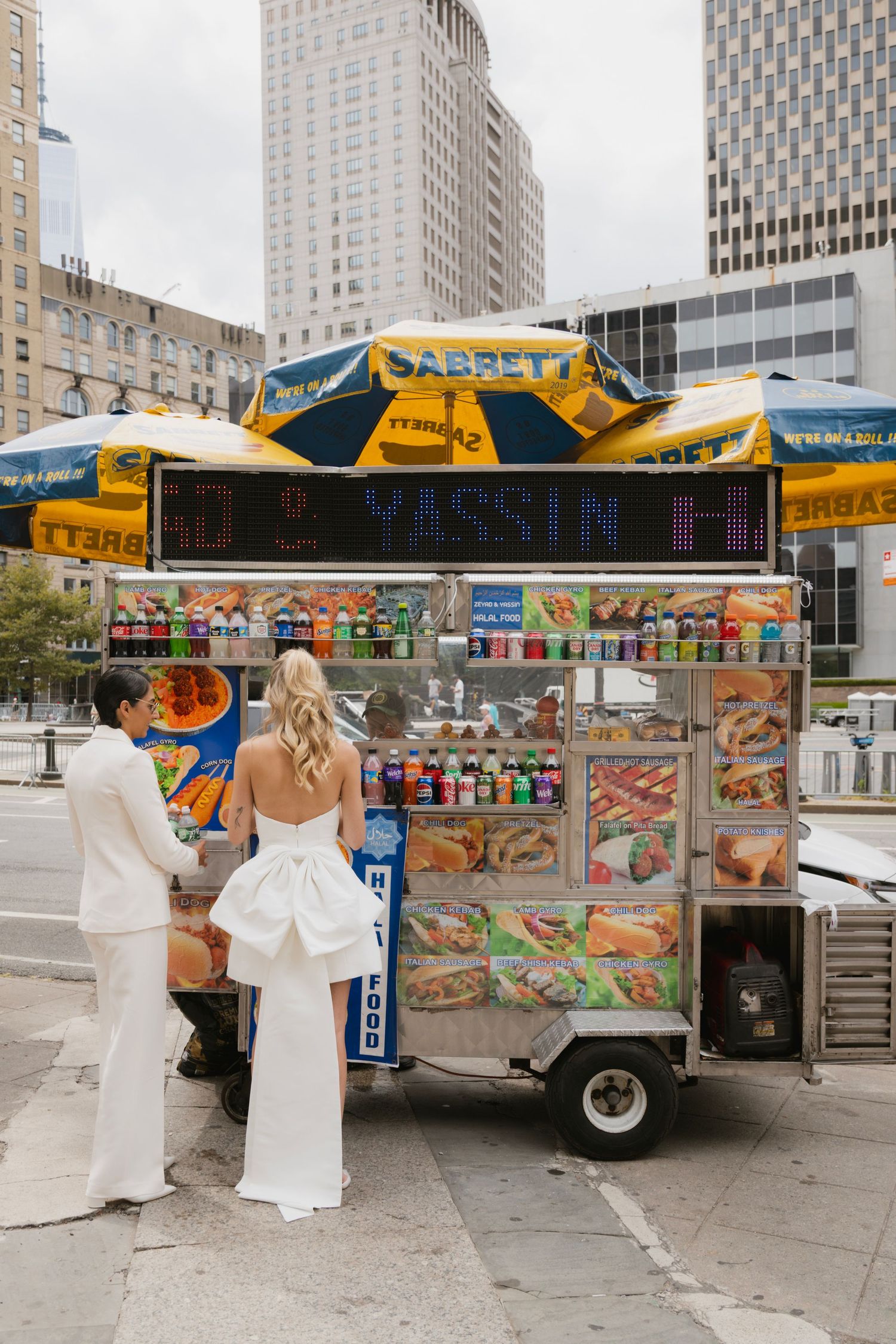 Food vendor cart with yellow umbrellas selling hot dogs and snacks on a city street with tall buildings in background.