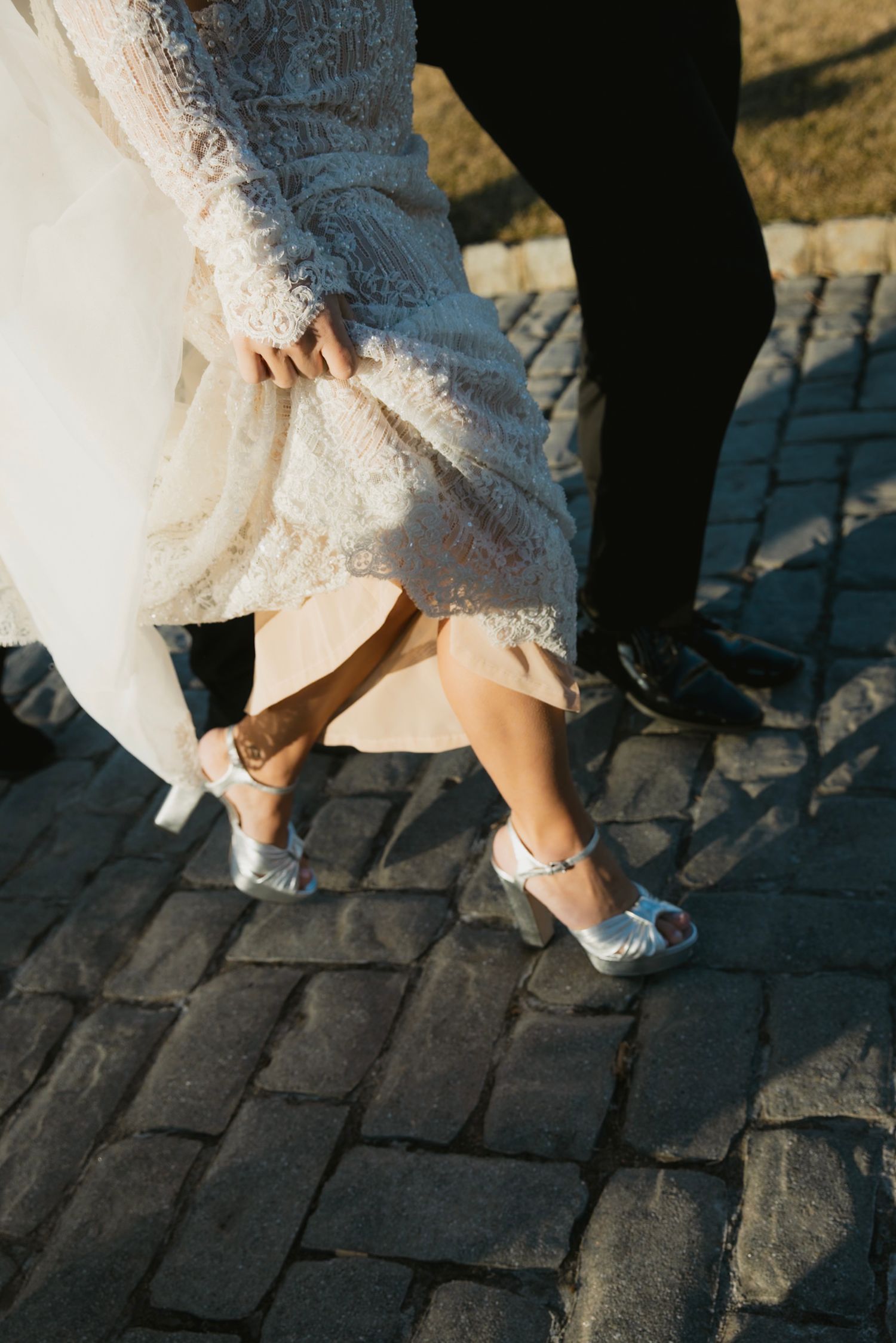 Close-up of bride's silver shoes and lace dress hem on cobblestone path.