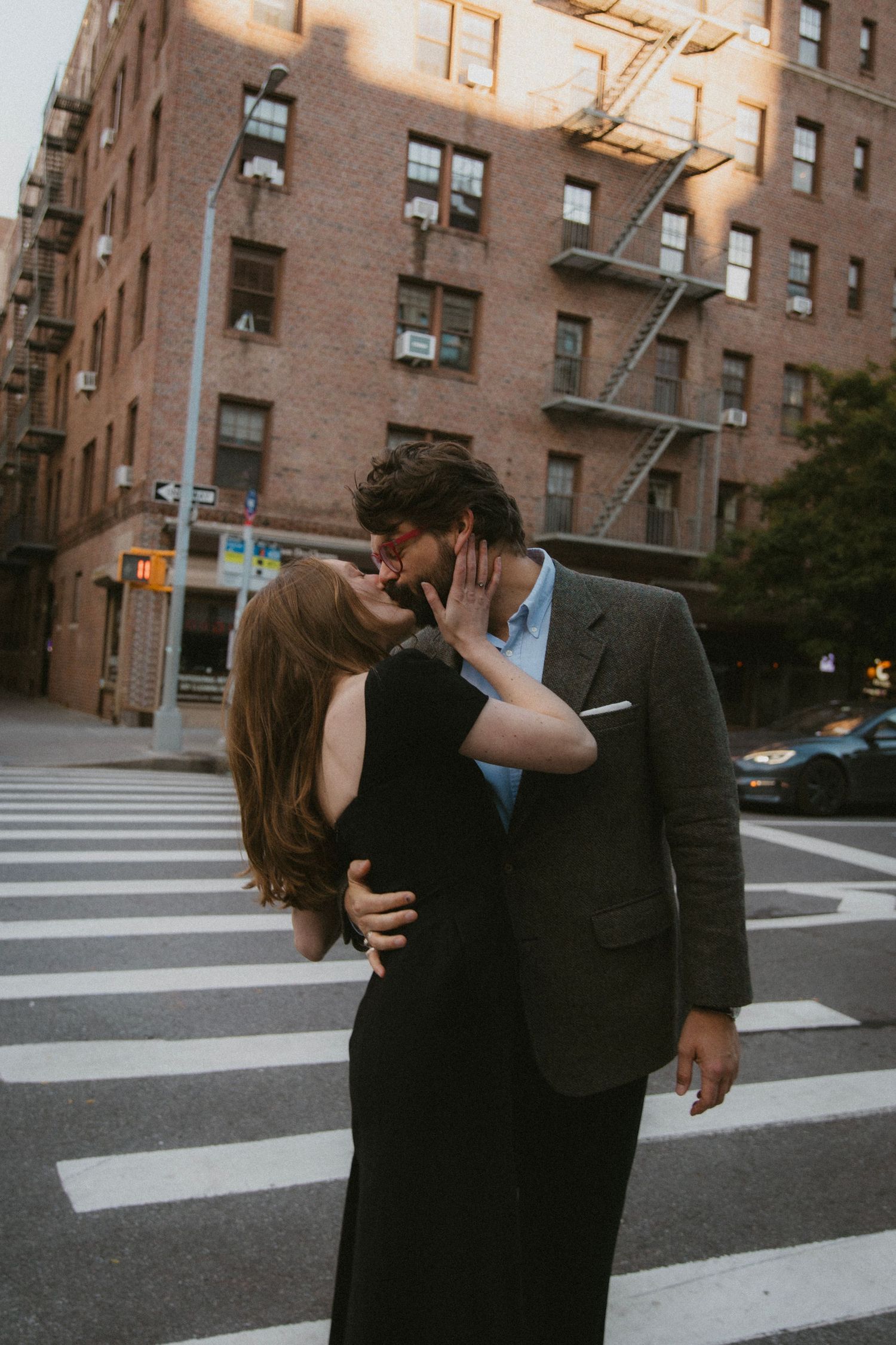A couple shares a romantic embrace on a city crosswalk with brick apartment buildings in the background at dusk.