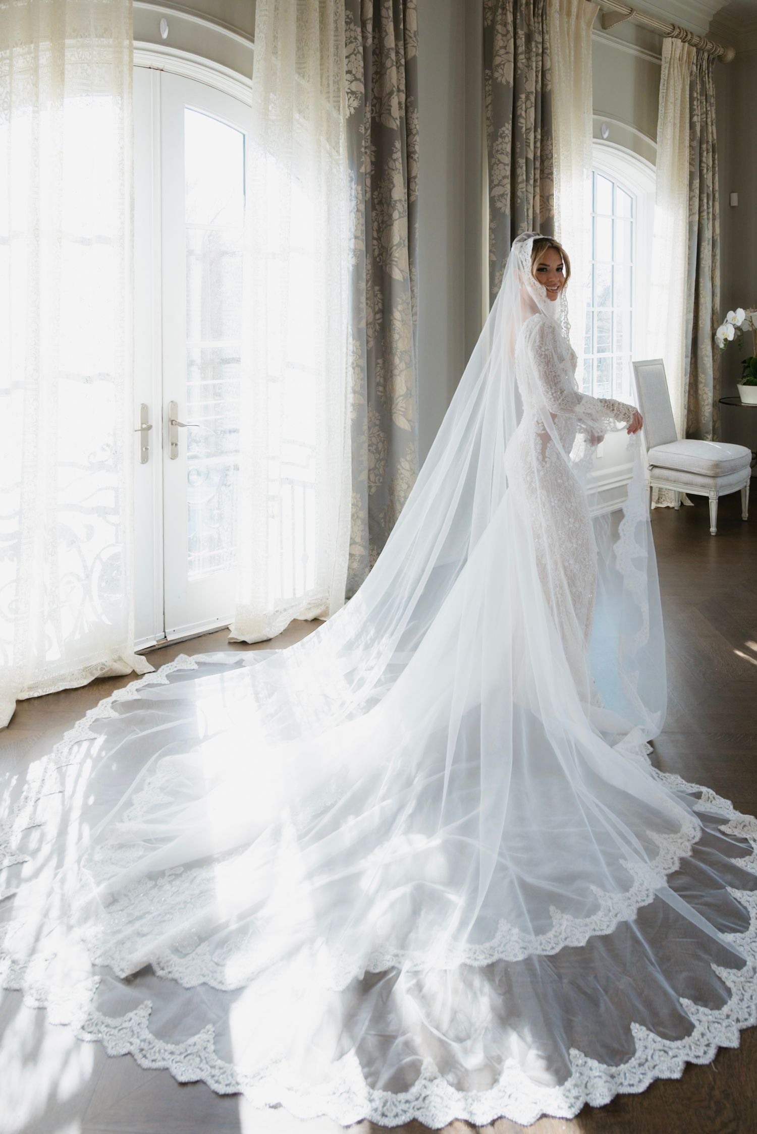 A flowing white wedding gown with long cathedral train swirls gracefully in natural window light.