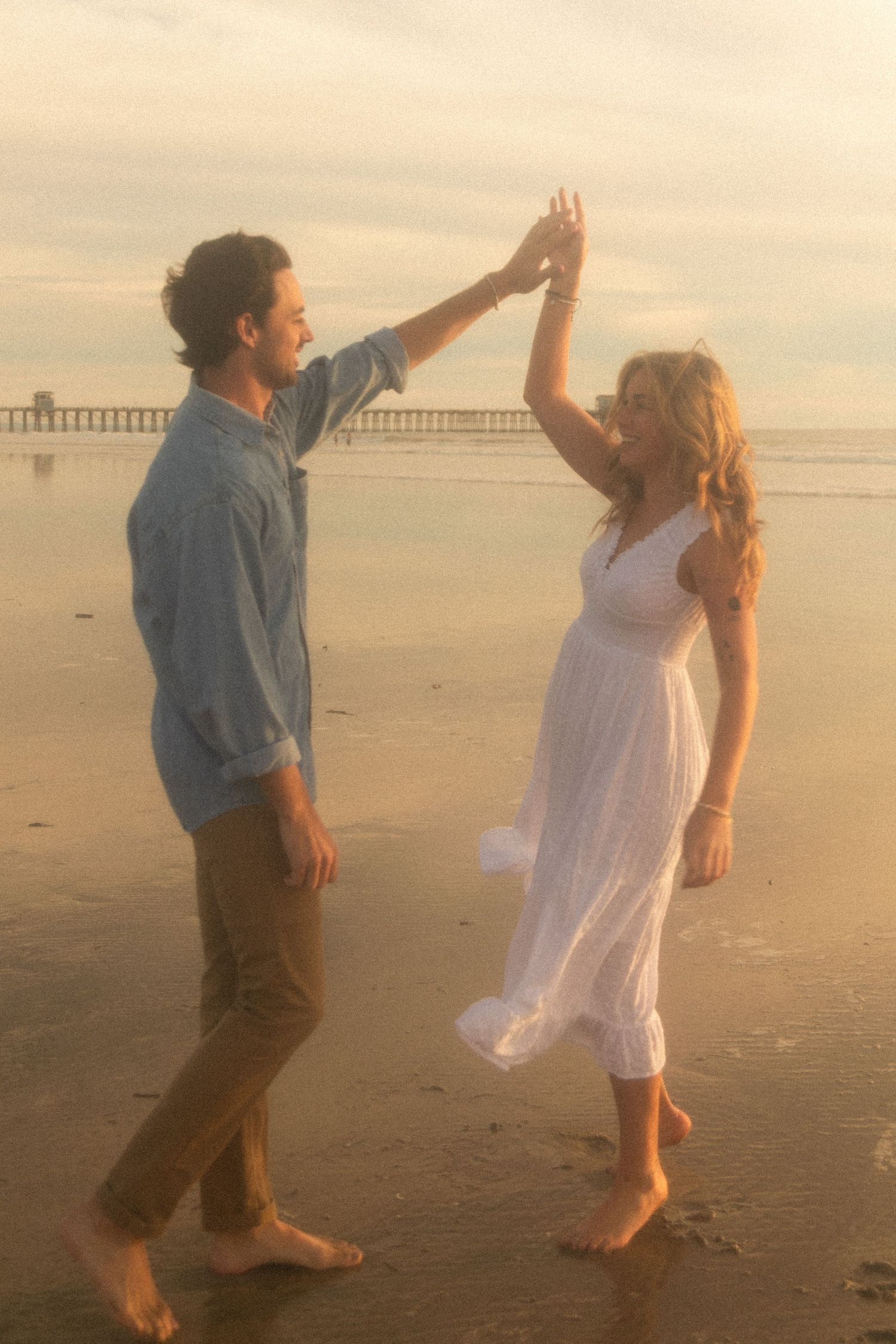 Two people in casual attire dance playfully on a misty beach at sunset with a pier visible in the distance.