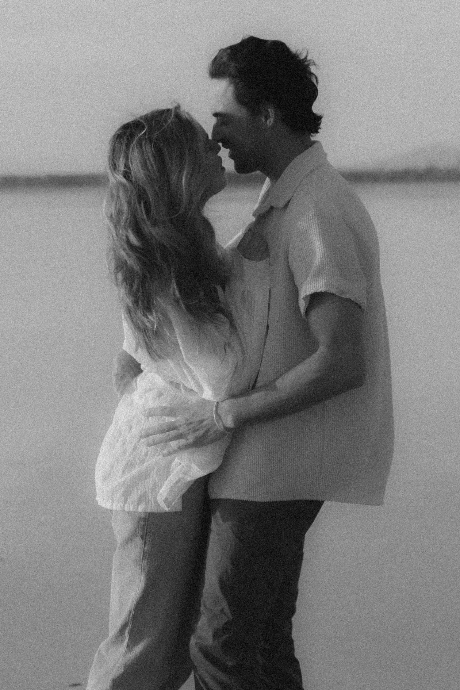 A black and white photo series captures intimate moments between a couple embracing on a misty beach.