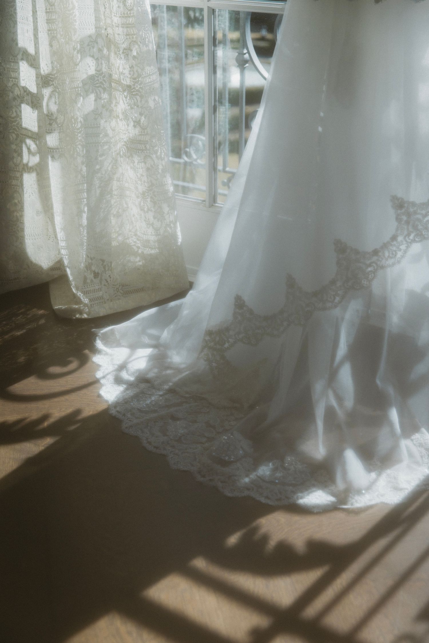 Delicate lace wedding dress hem with sunlight casting shadows through sheer curtains.