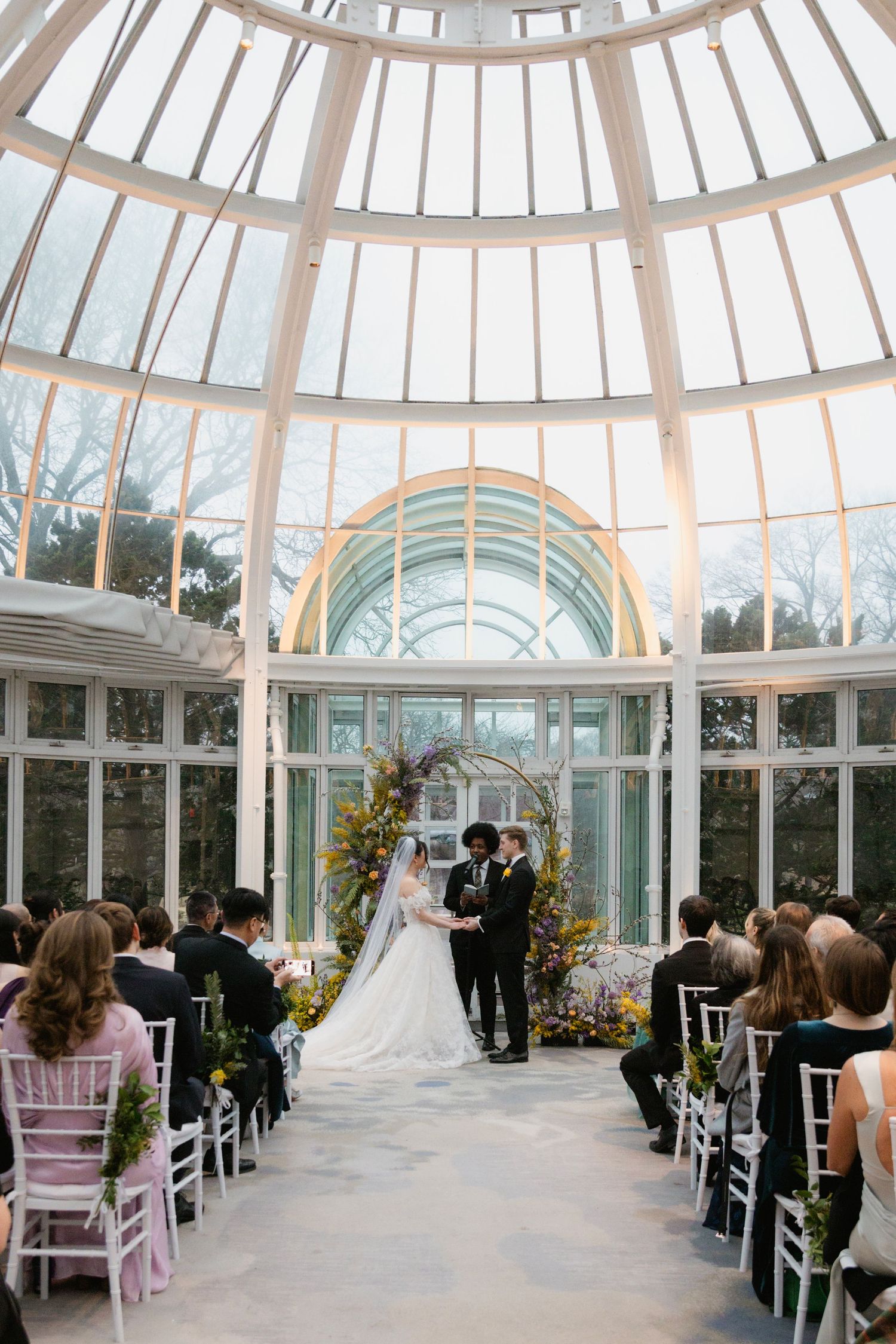 Wedding ceremony taking place in an elegant glass conservatory with domed ceiling and floral arch centerpiece.