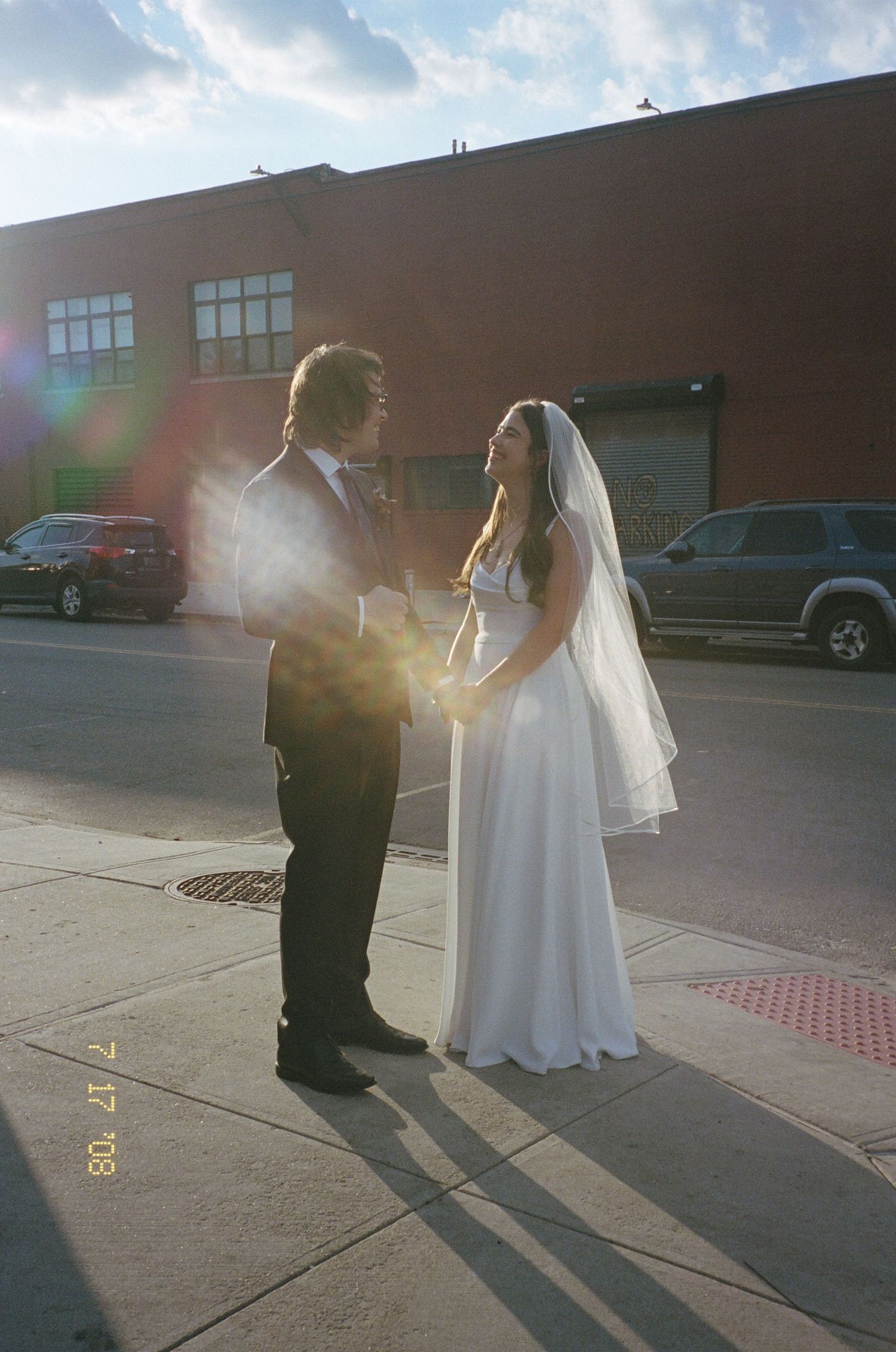 Wedding couple silhouetted against bright sunlight in urban parking lot setting.