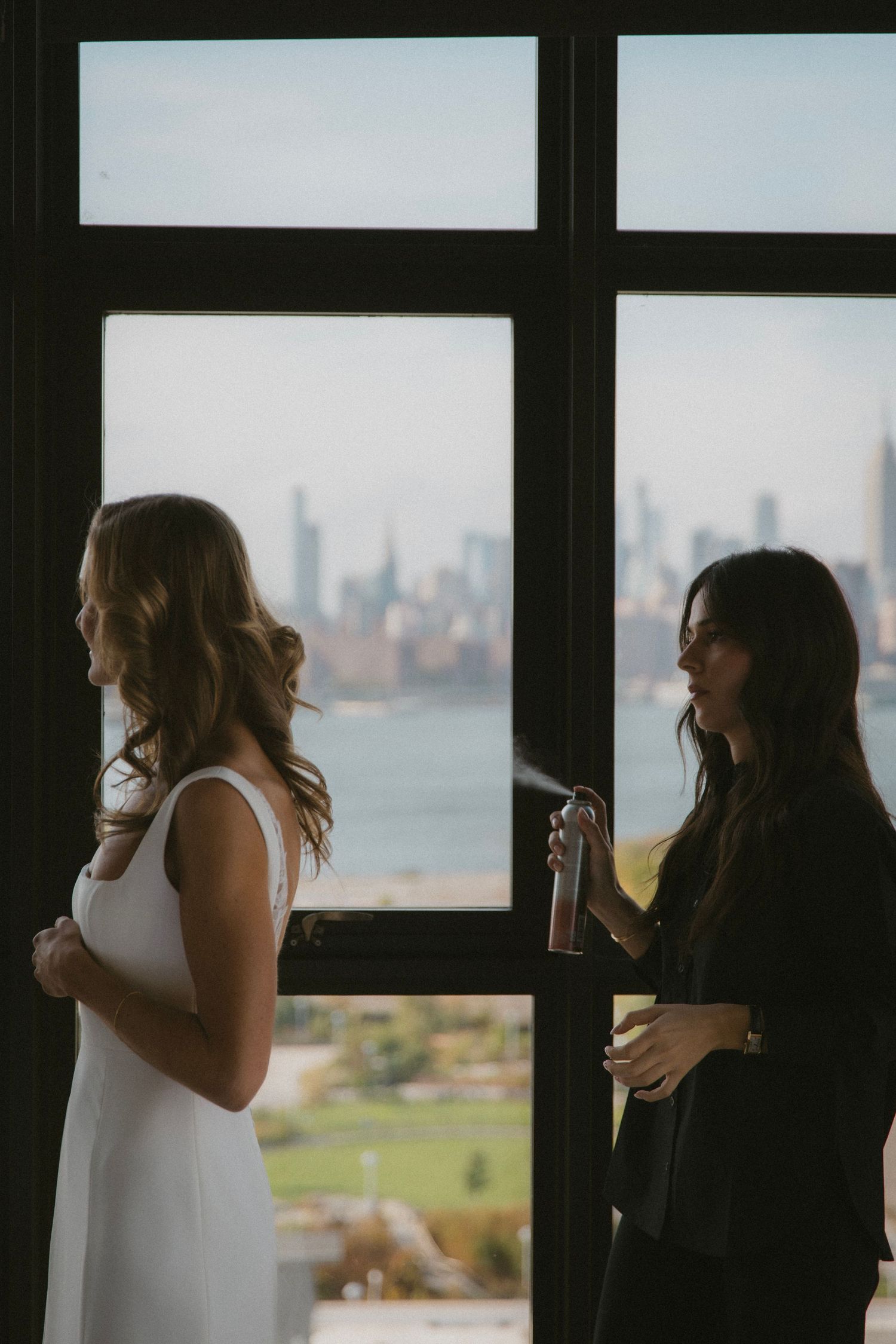 Two women in contrasting outfits stand by a window overlooking a city skyline.