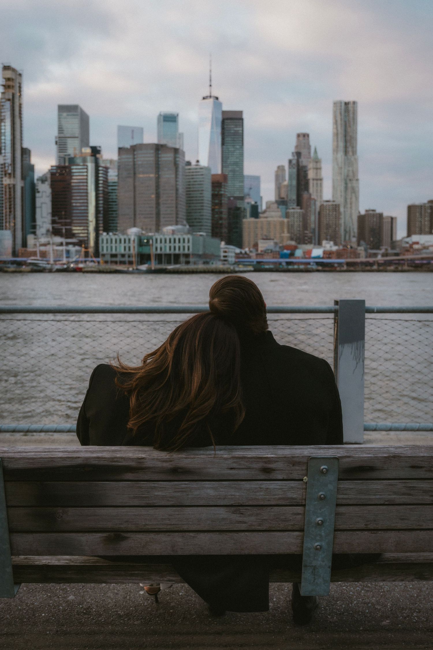 A person sits alone on a bench at the waterfront with the New York City skyline rising dramatically in the background.