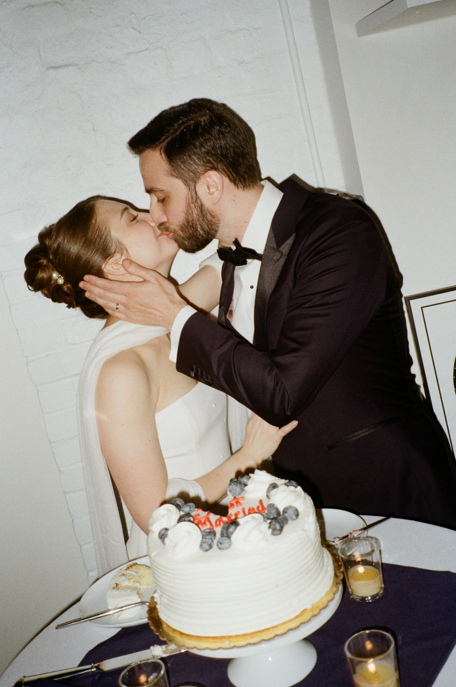 Newlyweds share a romantic kiss over their white wedding cake during the reception.