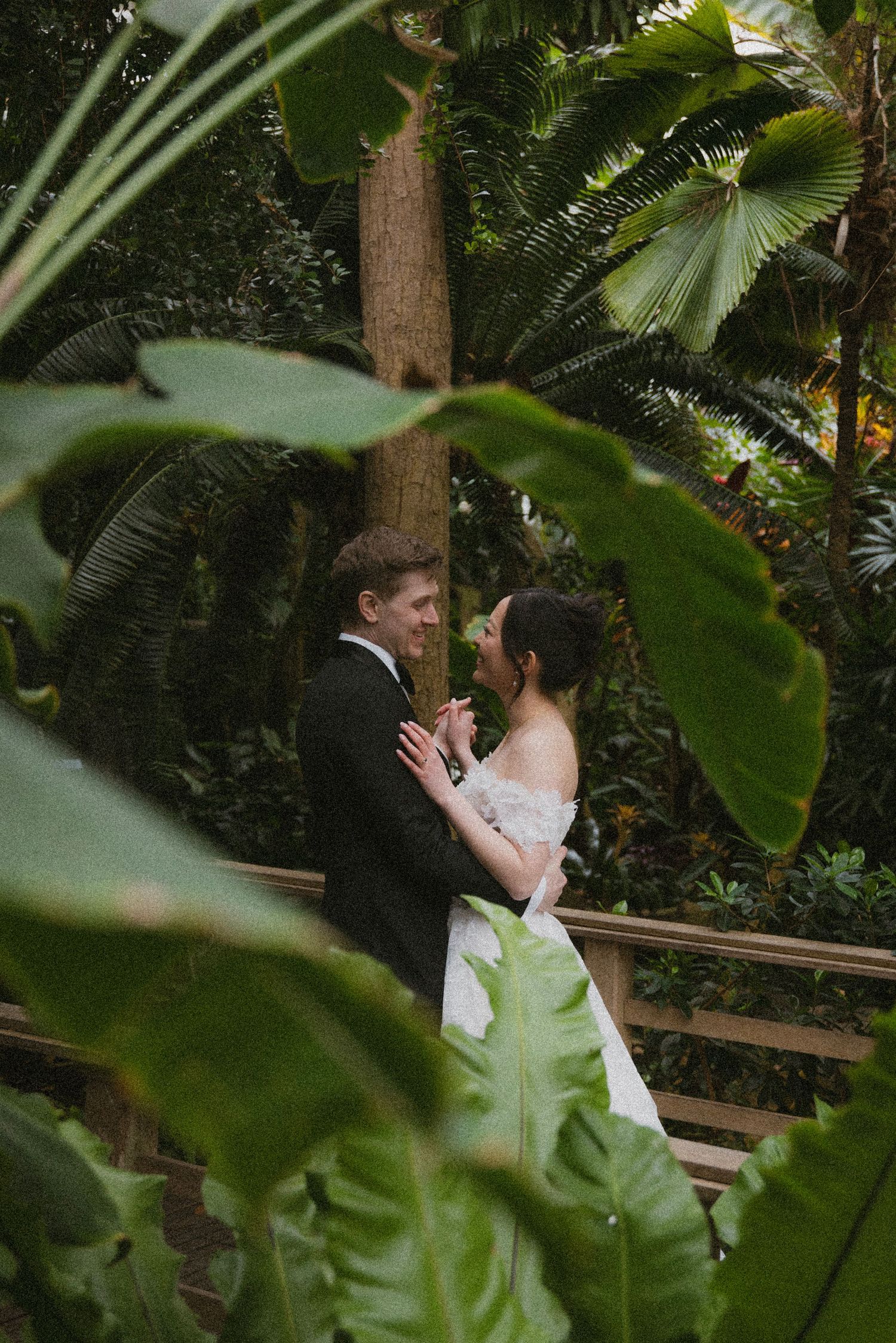 A romantic wedding couple embracing in a lush tropical garden setting with palm fronds and greenery framing the scene.