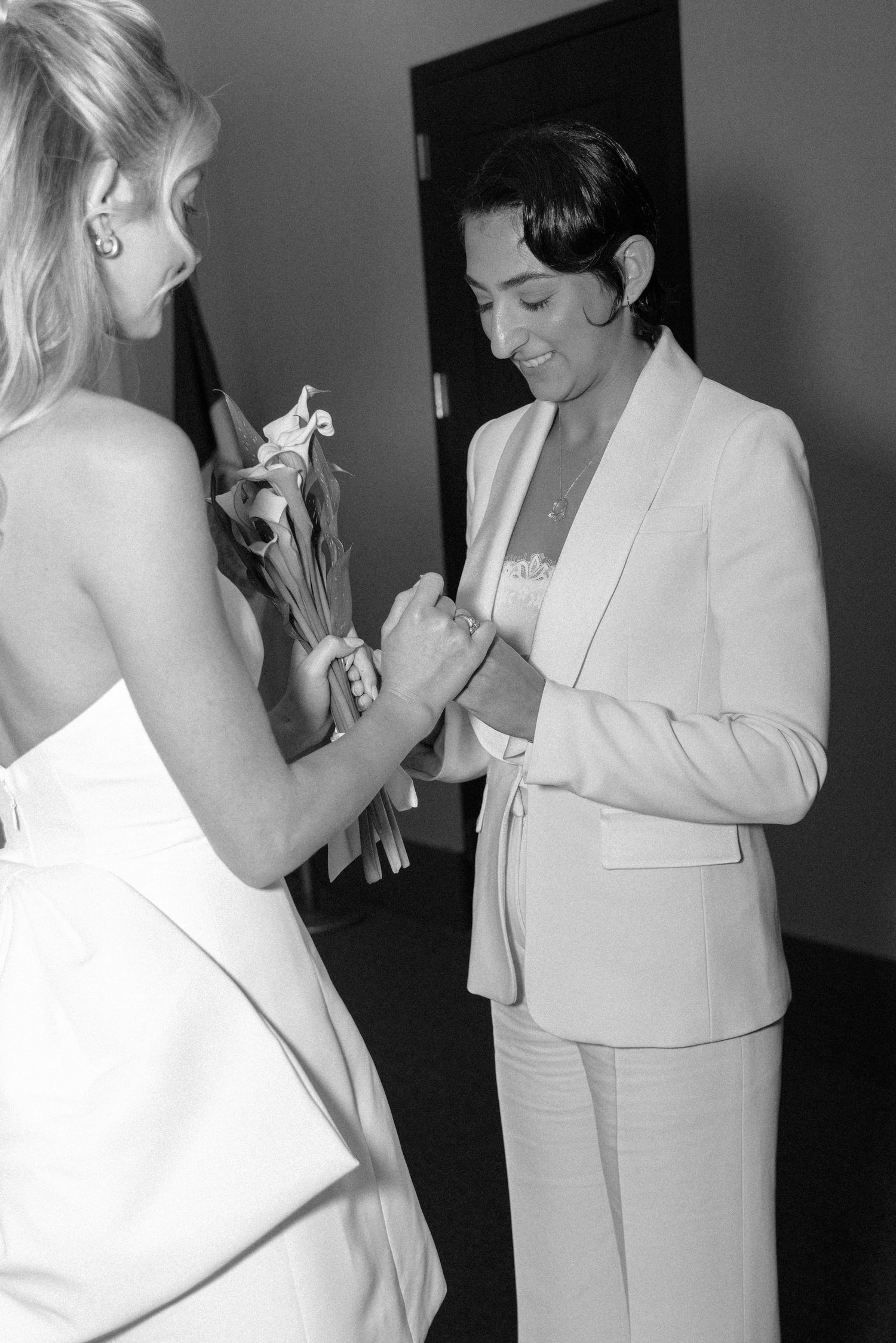 Two people in white formal attire interact during an event, with one holding flowers.