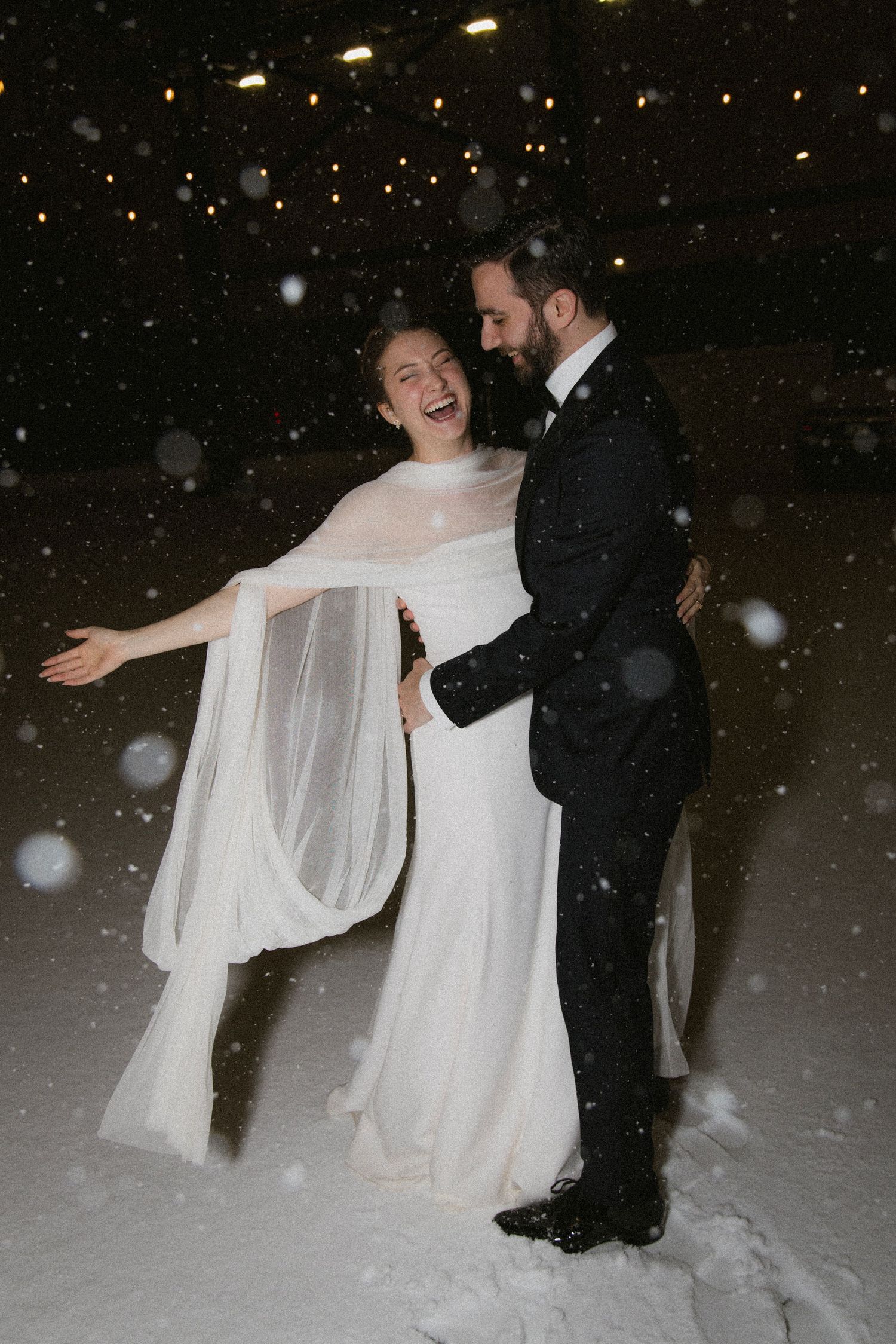 A bride and groom dance together in falling snow at night with twinkling lights in the background.