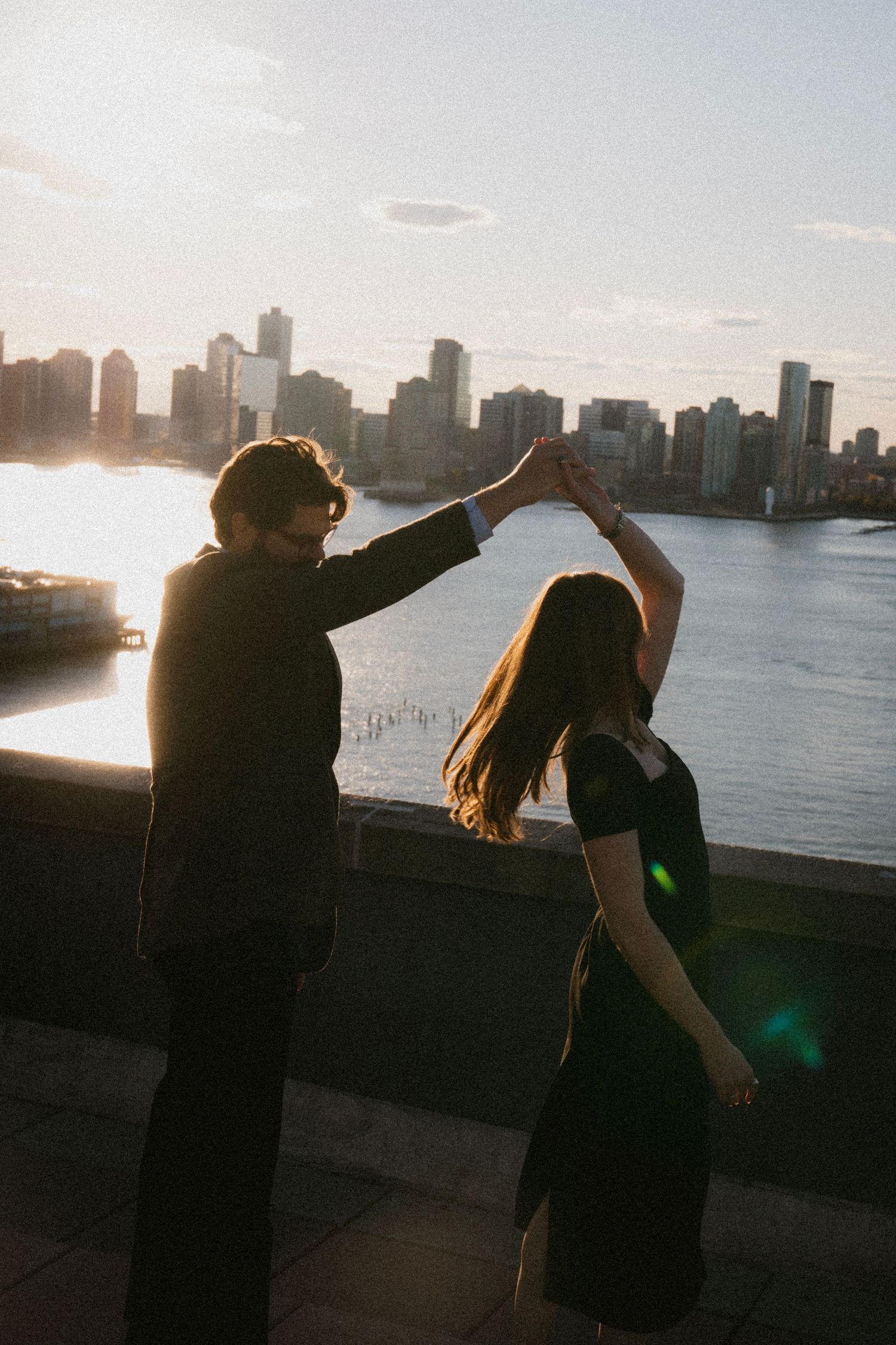 Two silhouettes dance together against a cityscape backdrop at sunset along the waterfront.