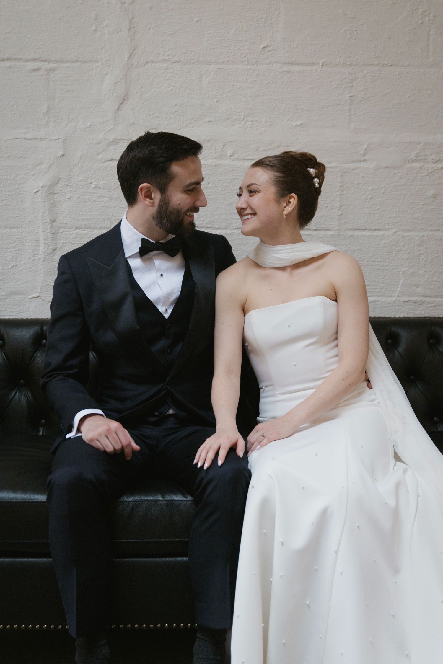 A tender moment between newlyweds as they sit on a black leather couch.