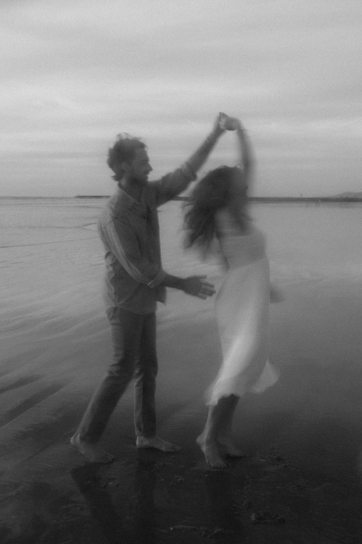 A couple dances together in silhouette on a beach at dusk, creating a romantic black and white scene by the water.
