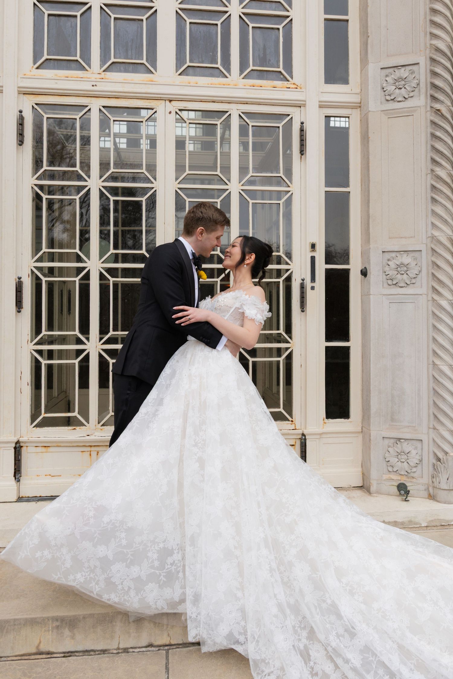 A romantic wedding couple embraces in front of a grand white mansion with elegant French windows and architectural details.