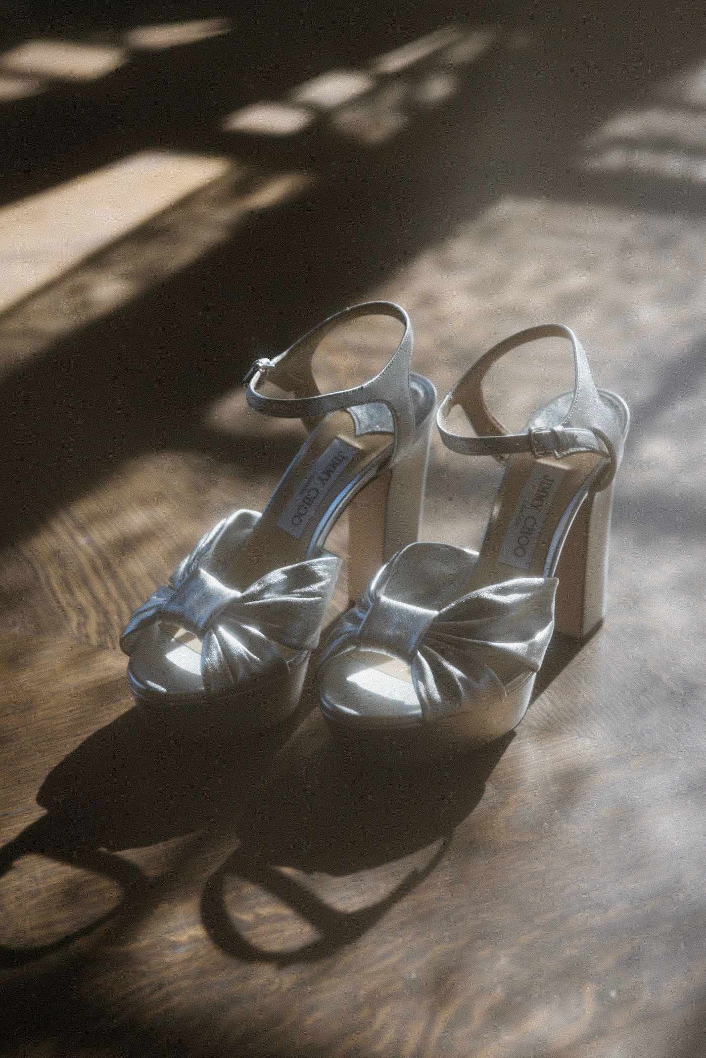 Metallic silver strappy high heel sandals photographed in dramatic lighting against a dark wooden surface.