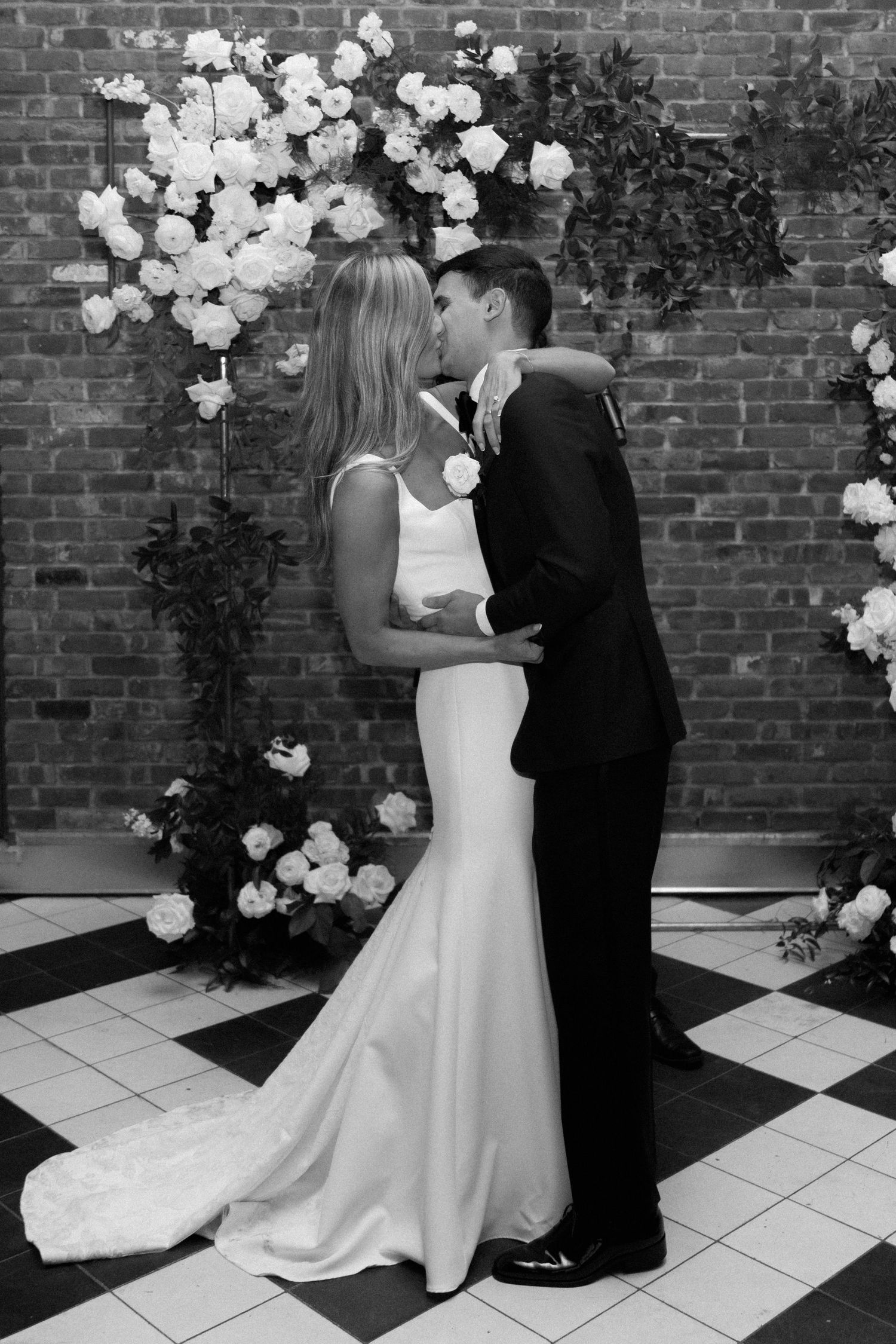 Black and white wedding photos against a brick wall decorated with white balloon arch and floral arrangements.