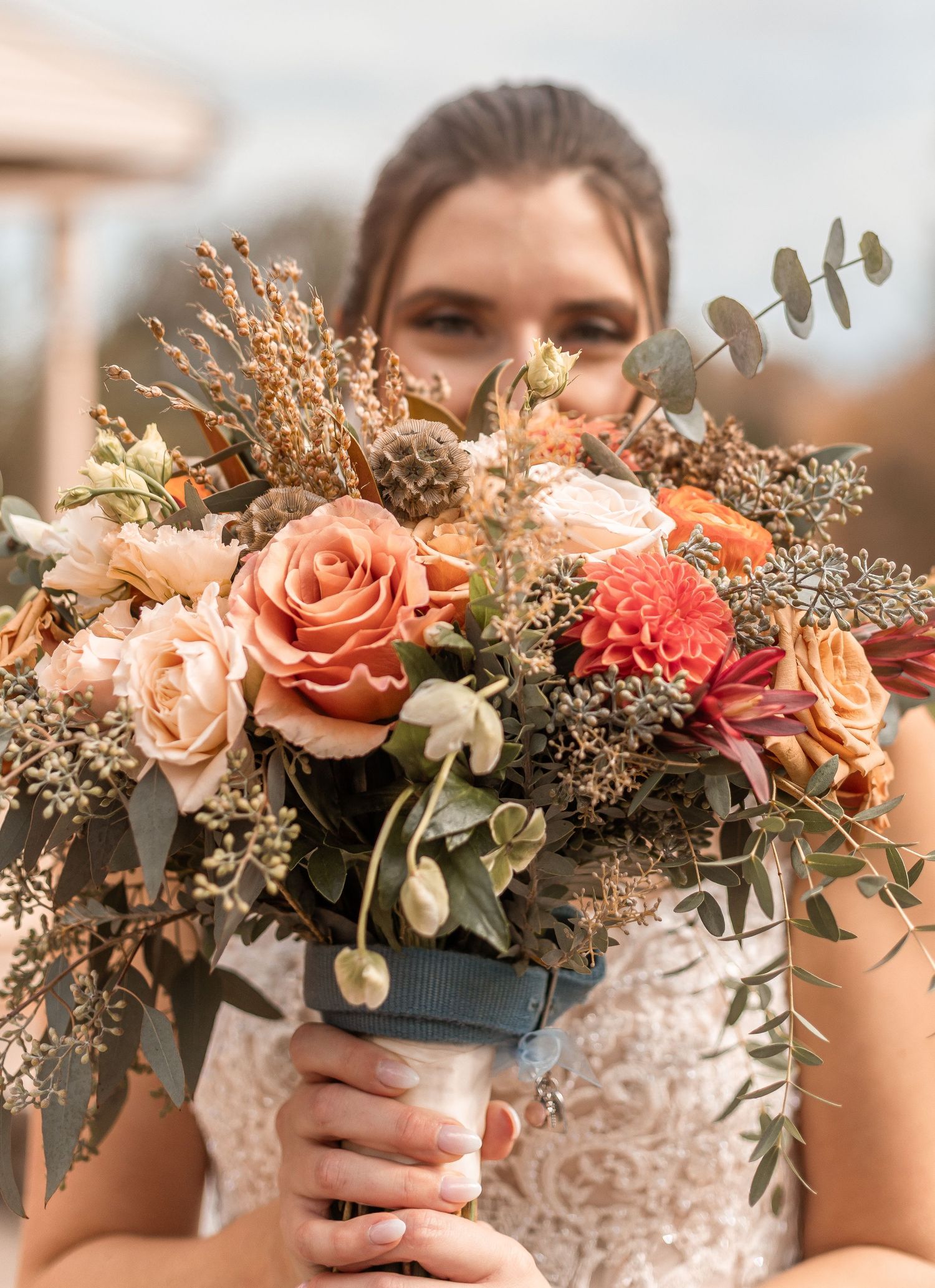 Bride holding a lush autumn bouquet with peach roses, dahlias, and eucalyptus in front of her face.
