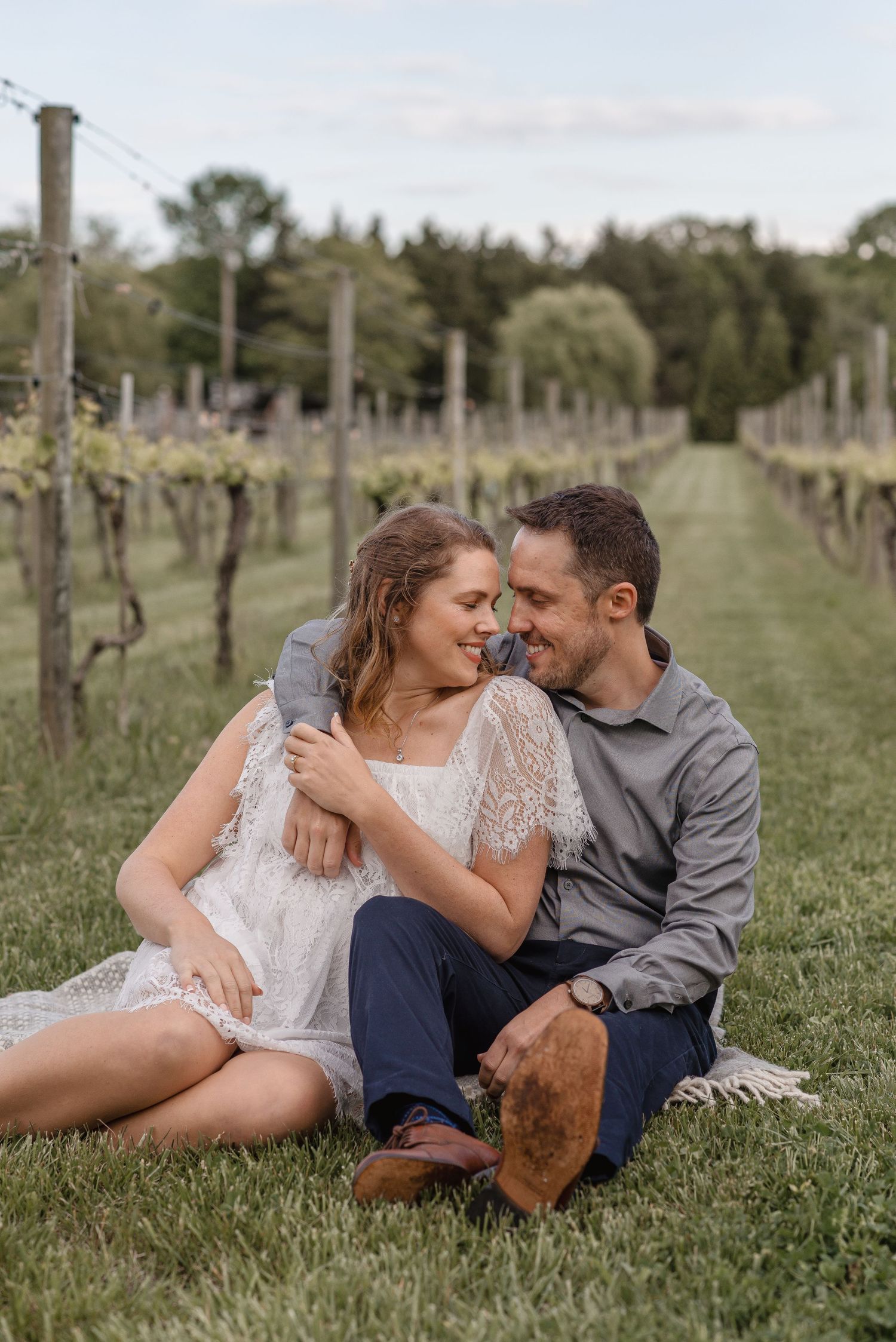 Couple sitting together on grass in a vineyard, smiling and embracing during an engagement session.