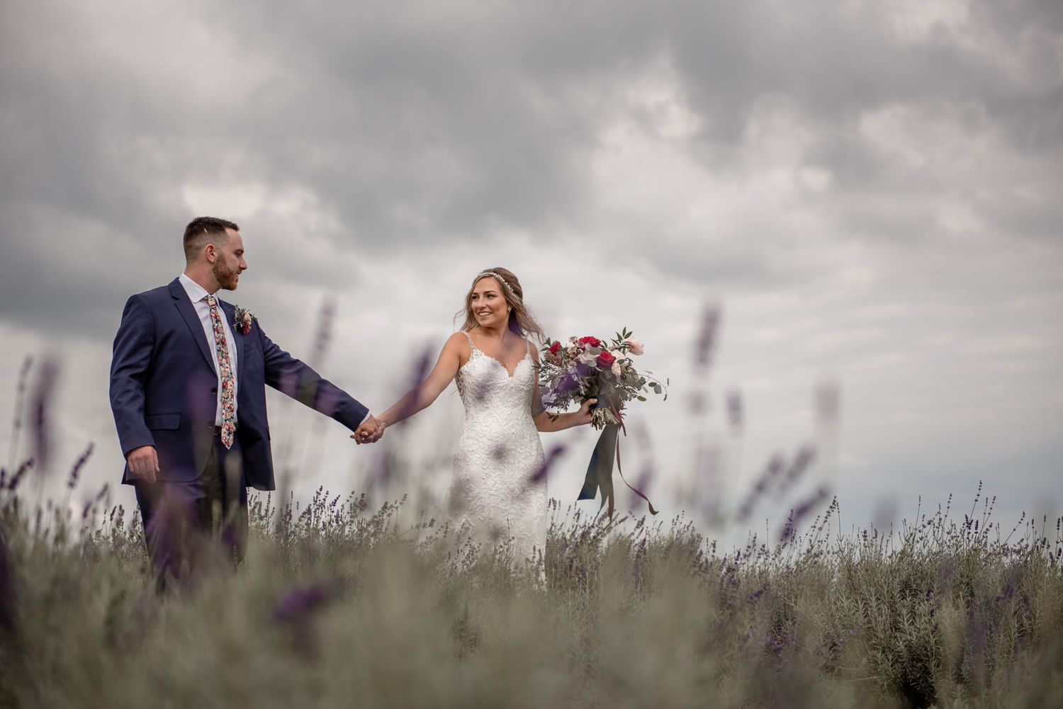 Bride and groom hold hands and smile while walking through a lavender field under a dramatic cloudy sky.