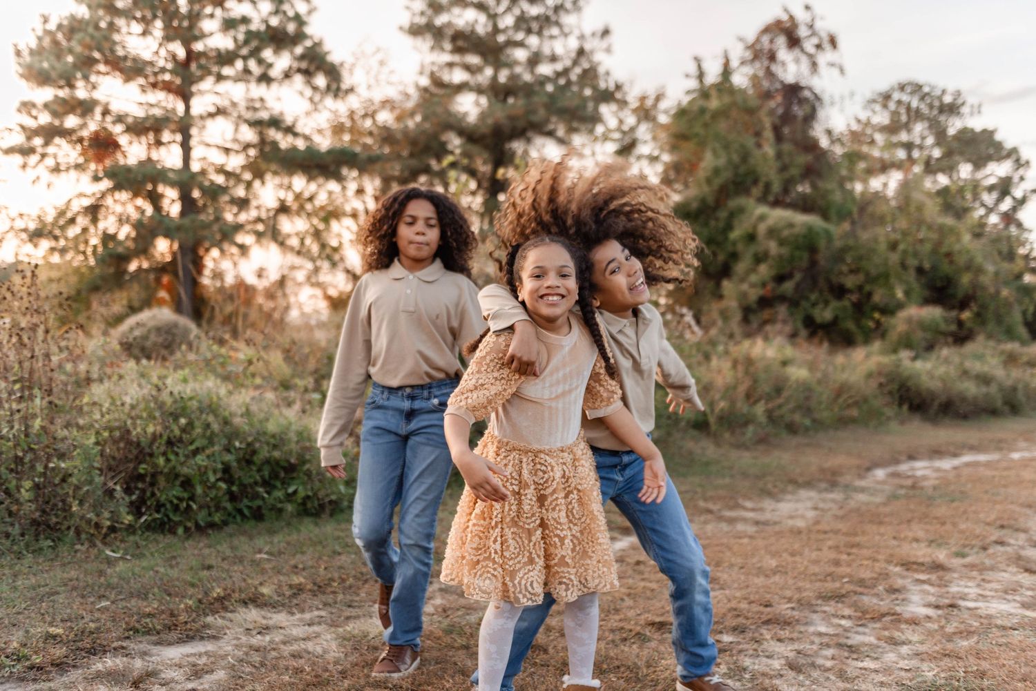 Three girls playfully dancing and laughing on a dirt path at golden hour outdoors.